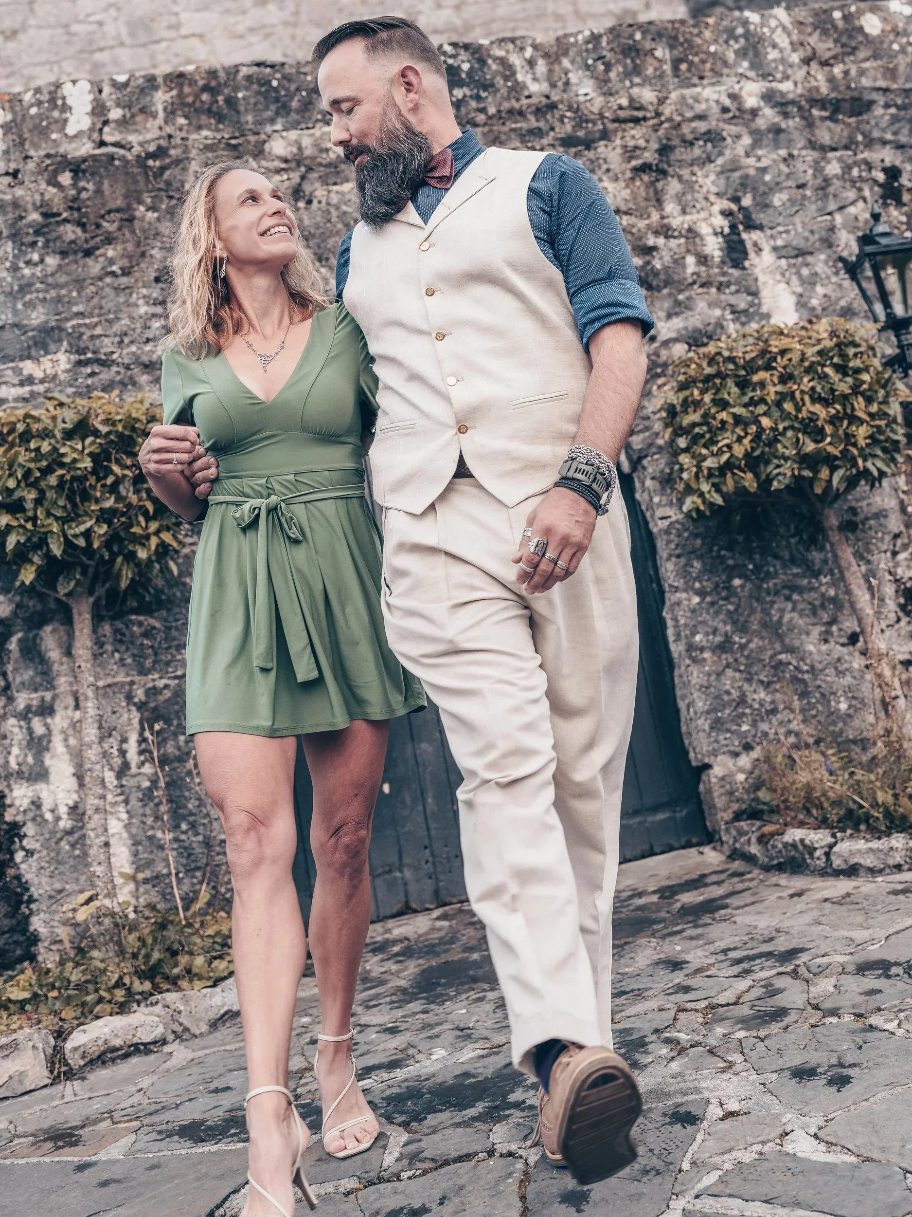 A man and woman holding each other and walking outdoors near a stone wall. The woman is wearing a green dress and high heels, and the man is dressed in a beige vest and pants with a blue shirt and bow tie.