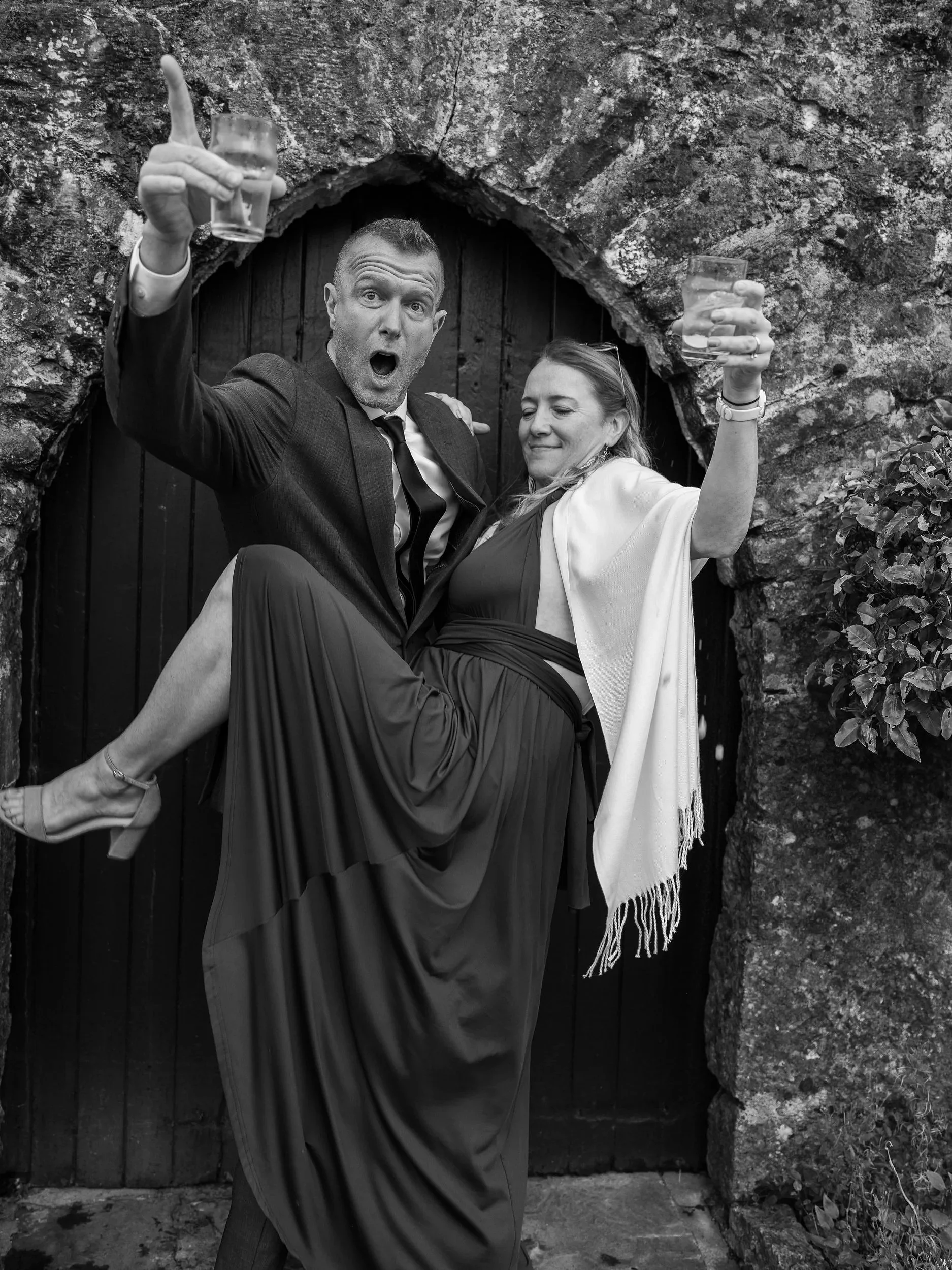 A man in a suit and a woman in a dress and shawl are posing in front of a stone archway, holding glasses of drink, with the man lifted off the ground and the woman smiling. Best-Fashion-Portrait-Commercial-Business-Headshots-Corporate- photography-Ma