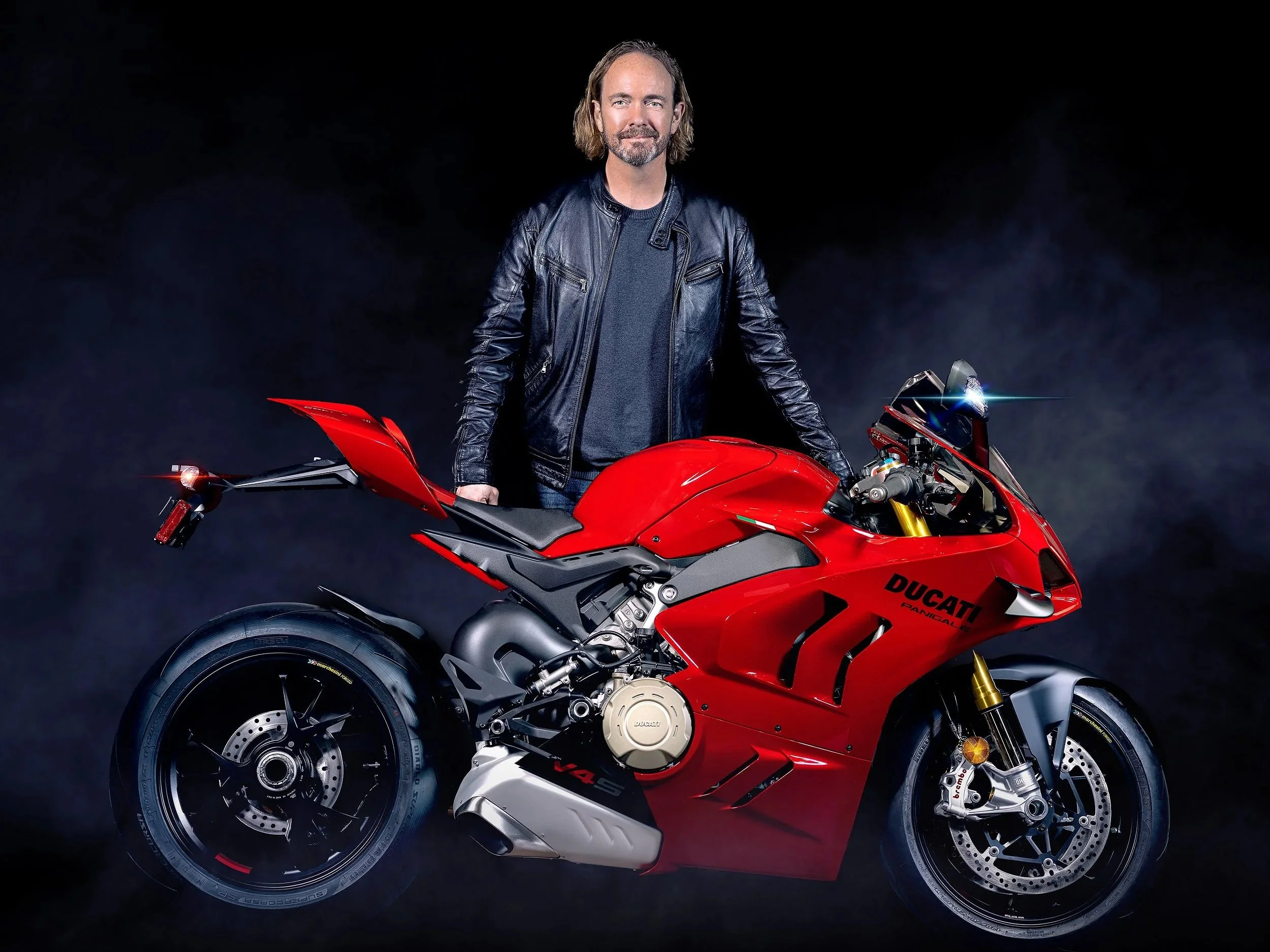 Man with shoulder-length hair and beard wearing a black leather jacket standing behind a red Ducati Panigale V4 motorcycle against a black background. Best-Fashion-Portrait-Commercial-Business-Headshots-Corporate- photography-Maryland-Washington D.C.