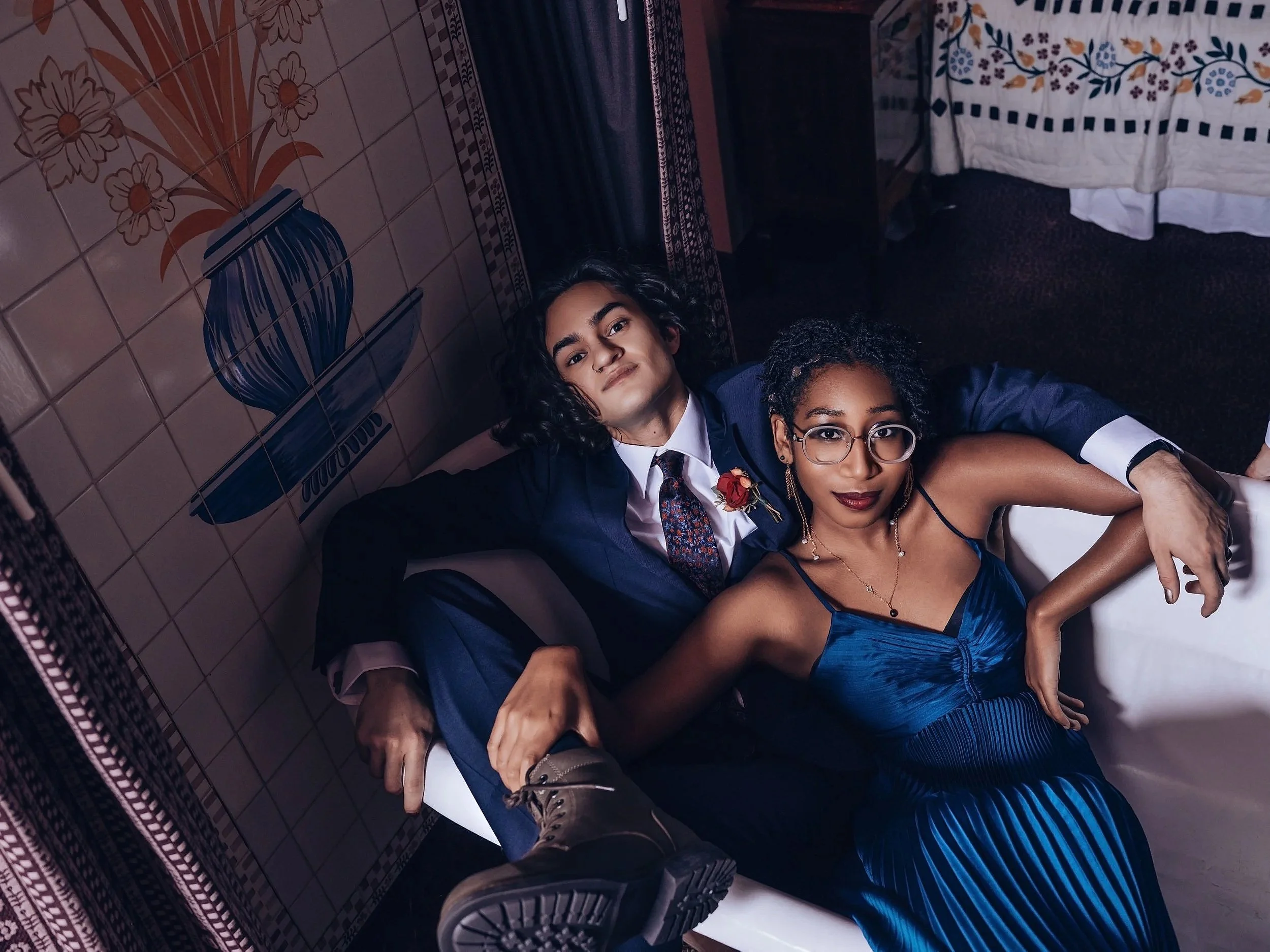 A young man and woman are lying on a bed in a room with colorful, decorative tile walls and a floral curtain, dressed in formal attire, with the man wearing a suit and the woman in a blue dress. Best-Fashion-Portrait-Commercial-Business-Headshots-Cor