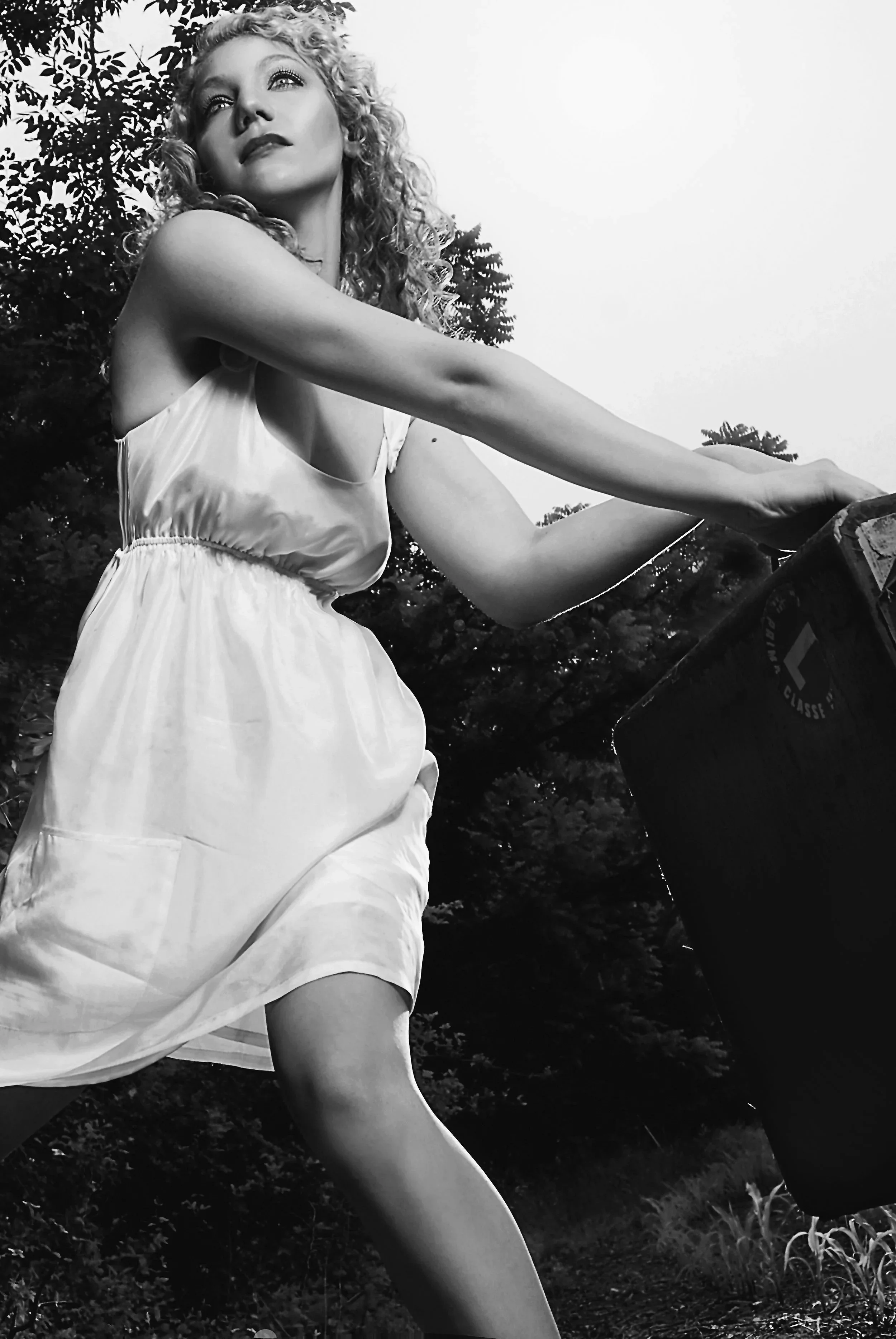 A woman with curly hair wearing a light-colored dress outdoors, reaching towards a trash bin, with trees in the background. Best-Fashion-Portrait-Commercial-Business-Headshots-Corporate- photography-Maryland-Washington D.C.-Pennsylvania-Virginia