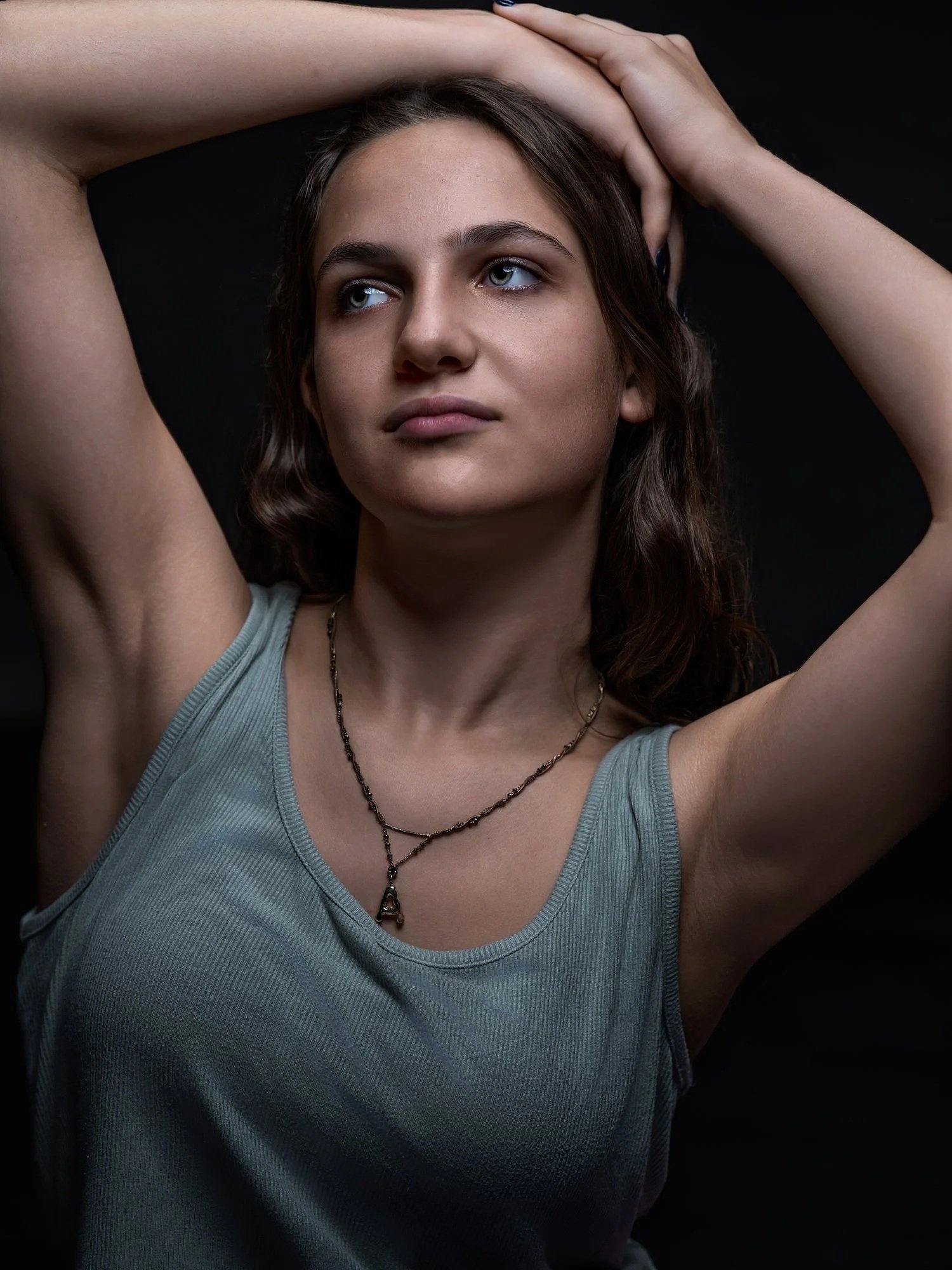 A young woman with blue eyes and brown hair, wearing a gray tank top and a necklace, posing with arms raised and hands behind her head against a black background. Best-Fashion-Portrait-Commercial-Business-Headshots-Corporate- photography-Maryland-Was