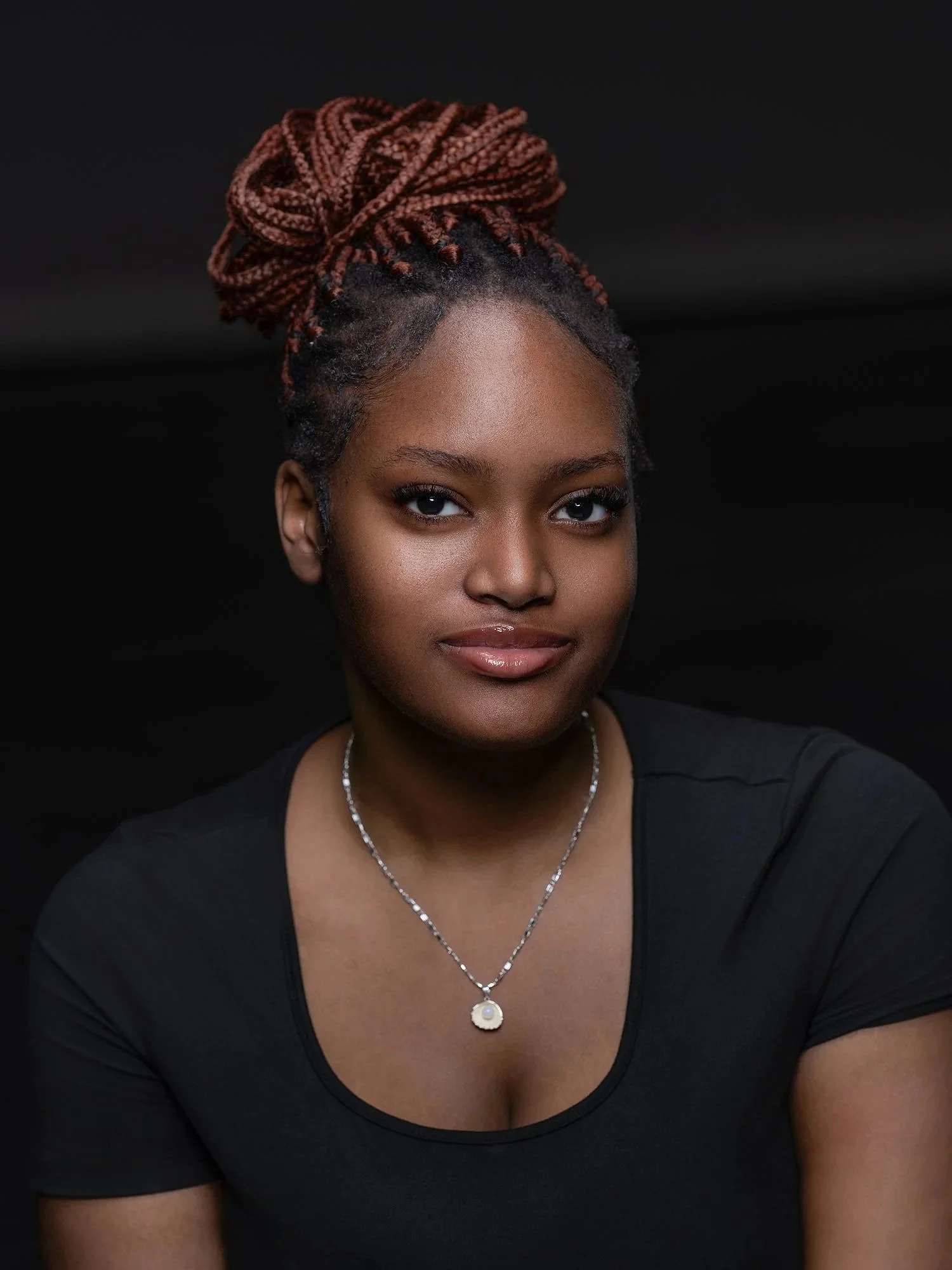 Portrait of a young African American woman with braided hair styled in an updo, wearing a black top and a silver necklace, against a dark background. Best-Fashion-Portrait-Commercial-Business-Headshots-Corporate- photography-Maryland-Washington D.C.-