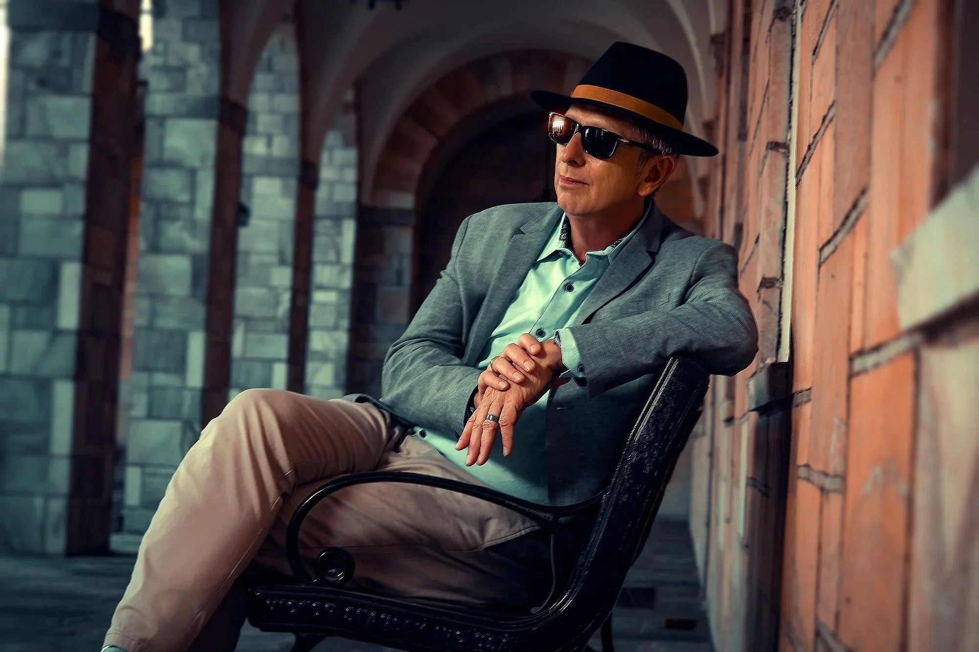 A stylish middle-aged man in sunglasses and a fedora hat, wearing a gray blazer and mint green shirt, sitting on a black metal outdoor chair against a brick wall with arched architecture in the background. Best-Fashion-Portrait-Commercial-Business-He