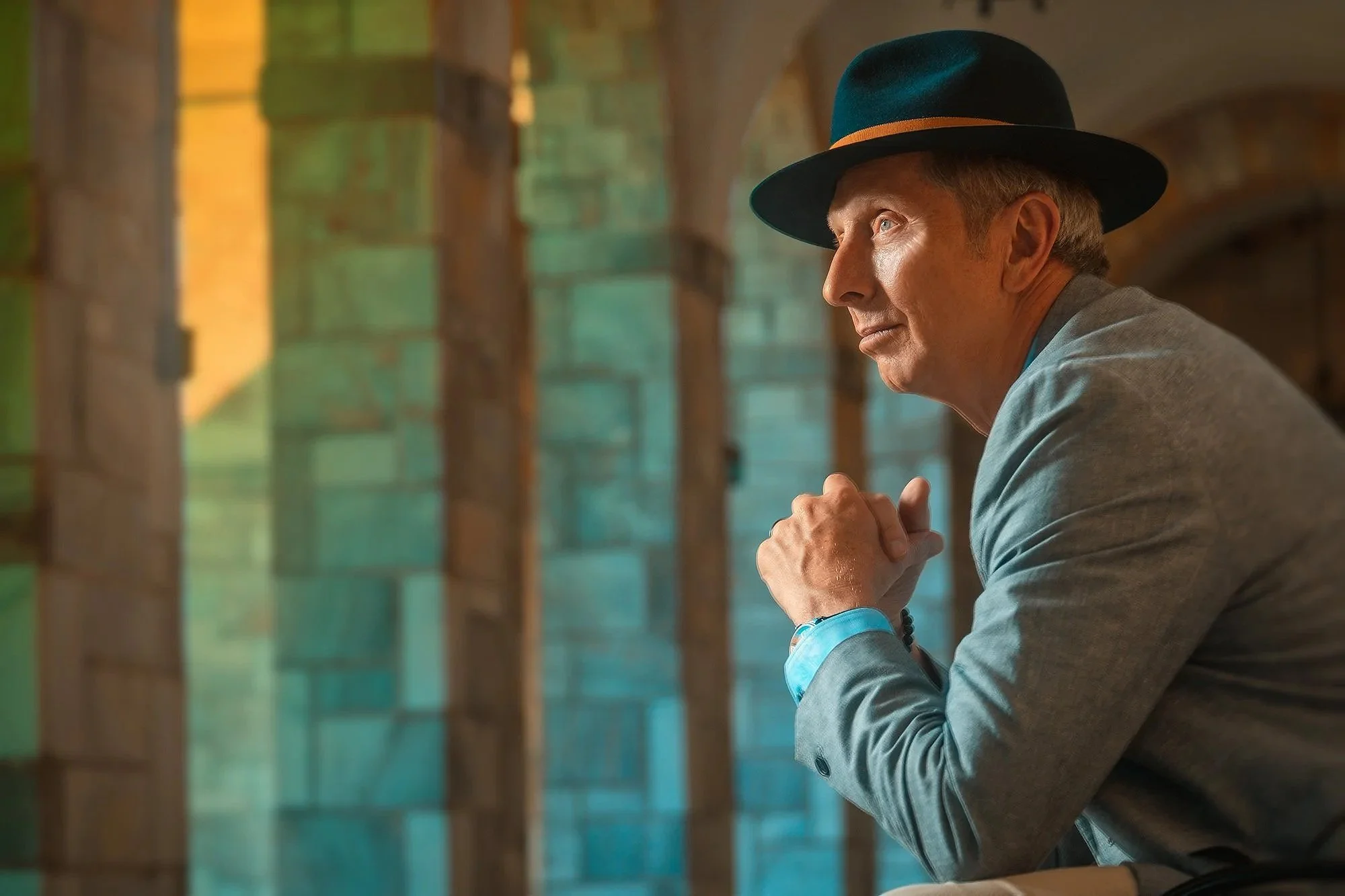 A man in a gray suit and teal hat sitting with hands clasped, looking thoughtfully to the right, sitting indoors with colorful brick walls and arches in the background. Best-Fashion-Portrait-Commercial-Business-Headshots-Corporate- photography-Maryla