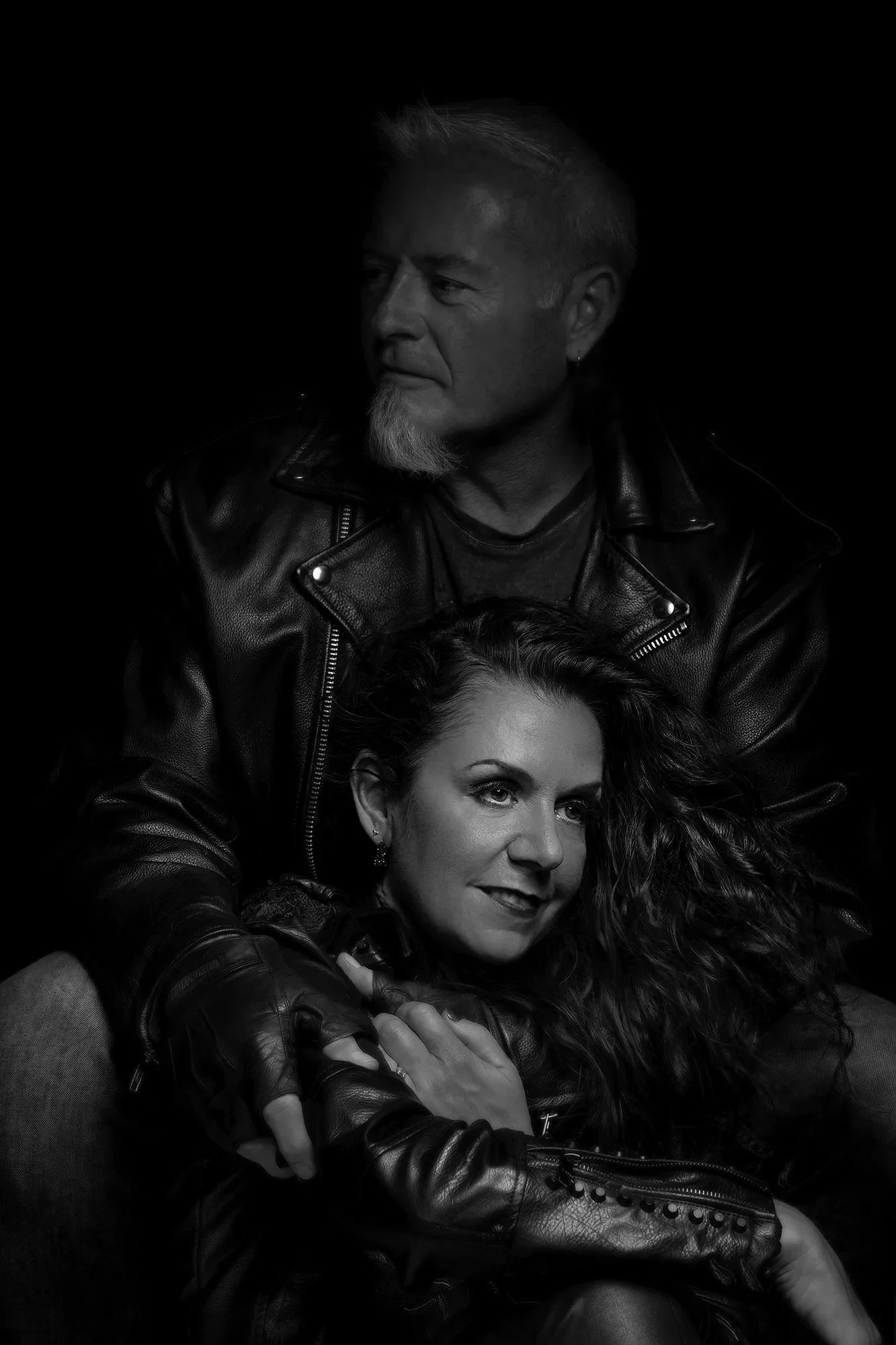 A black-and-white portrait of a man and woman, both wearing leather jackets, with the man standing behind and slightly above the woman who is sitting, both looking away from the camera.