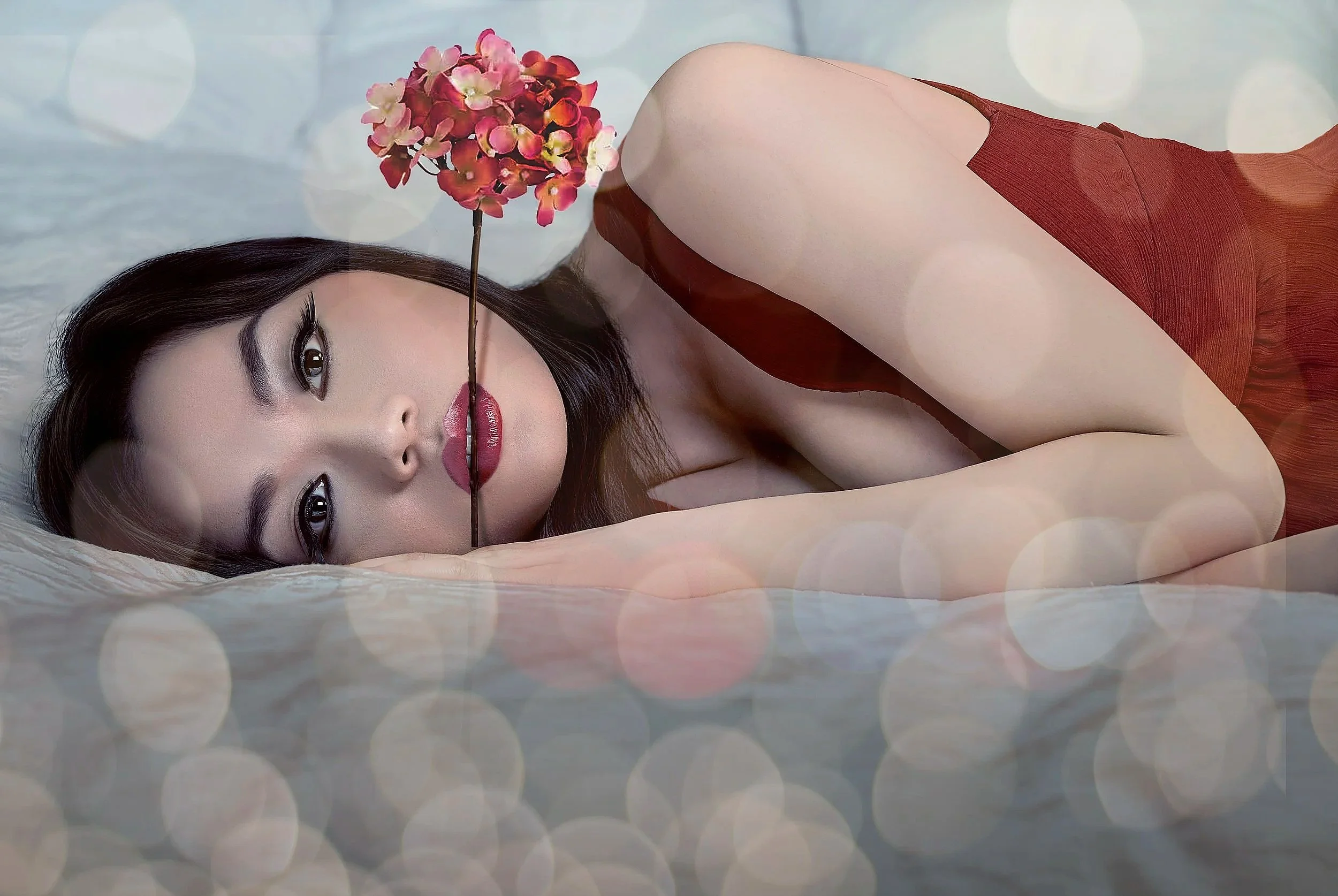 A woman with long dark hair lying on a bed, holding a flower stem with pink flowers between her lips, wearing a red dress, with soft blurred lights in the background.