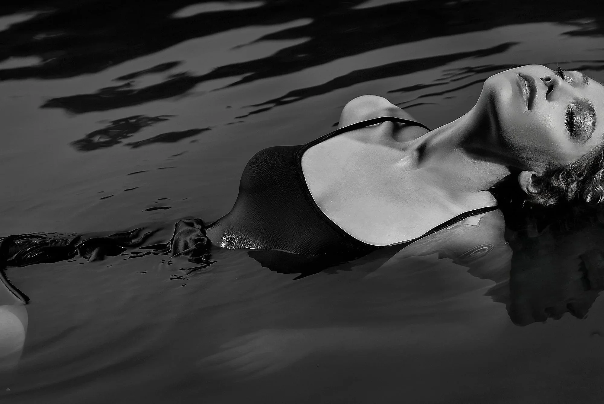 A woman in a black swimsuit floating on her back in water with eyes closed and head tilted back, black and white photograph.