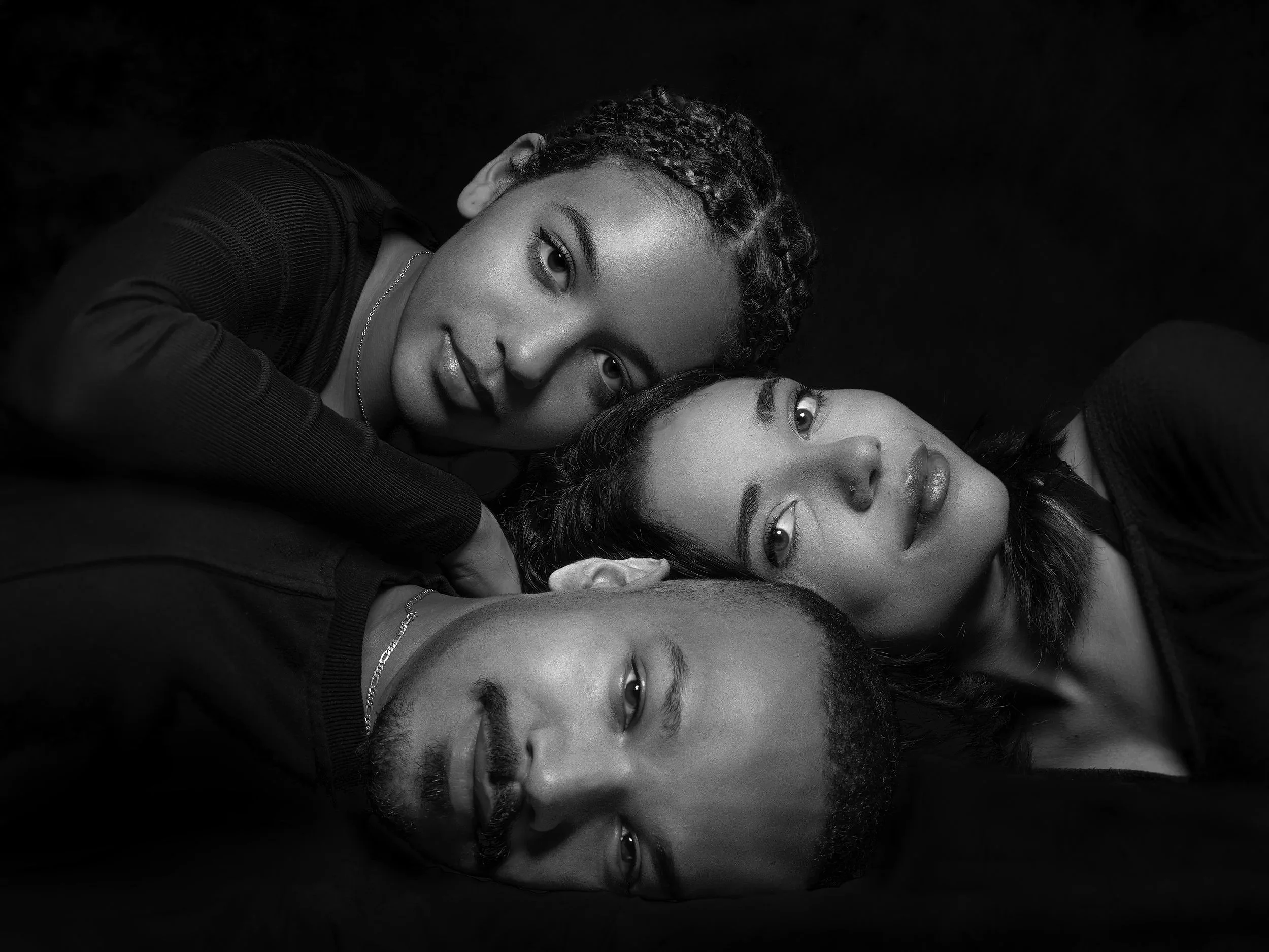 Black and white photo of three young adults lying on a dark surface, their heads close together, looking at the camera.