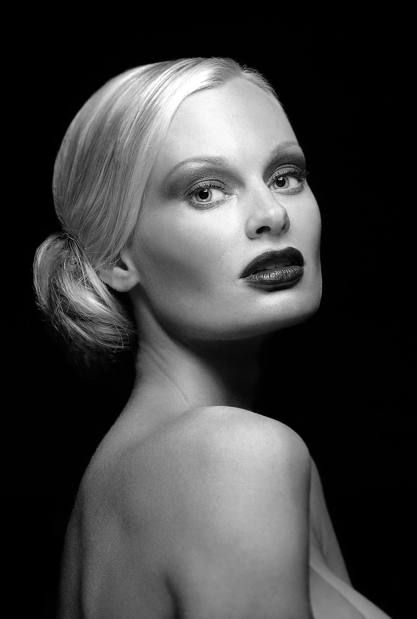 Black and white portrait of a woman with platinum blonde hair styled in a low side bun, wearing makeup with dark lipstick, looking over her shoulder against a dark background.