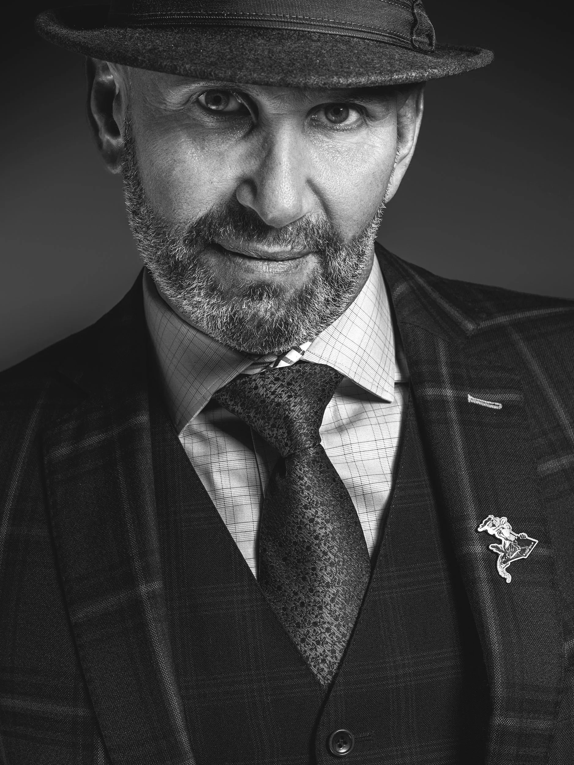 Black and white portrait of a man wearing a fedora, patterned tie, checkered dress shirt, and plaid blazer with a lapel pin featuring a horse and rider.