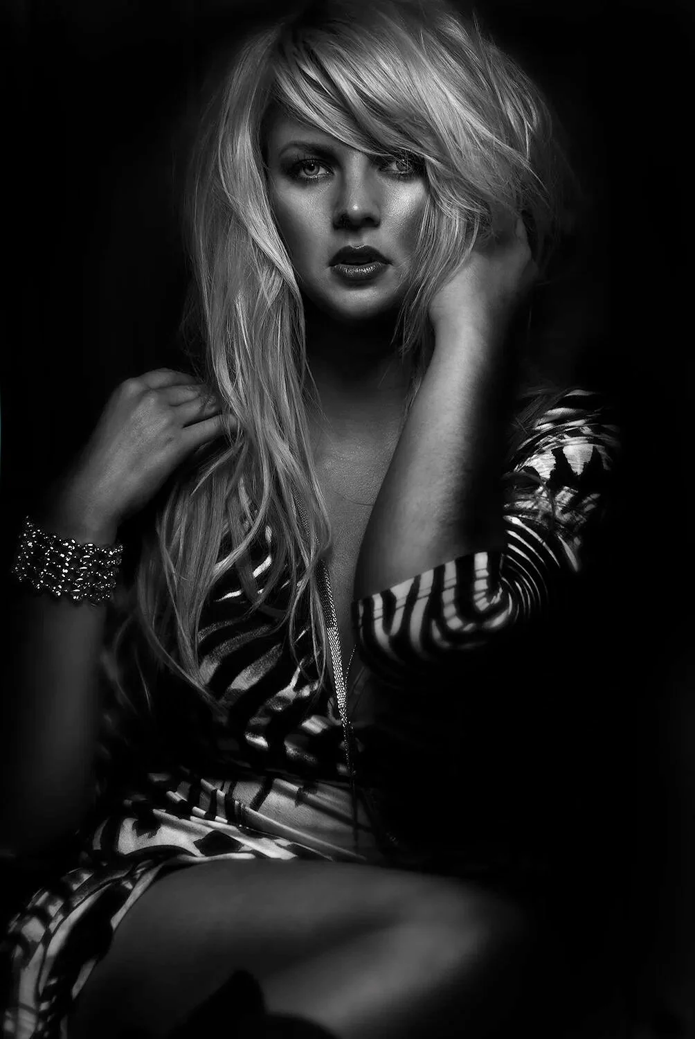 A black and white portrait of a woman with long, tousled blonde hair, wearing a patterned top and bracelet, looking directly at the camera with an intense expression.