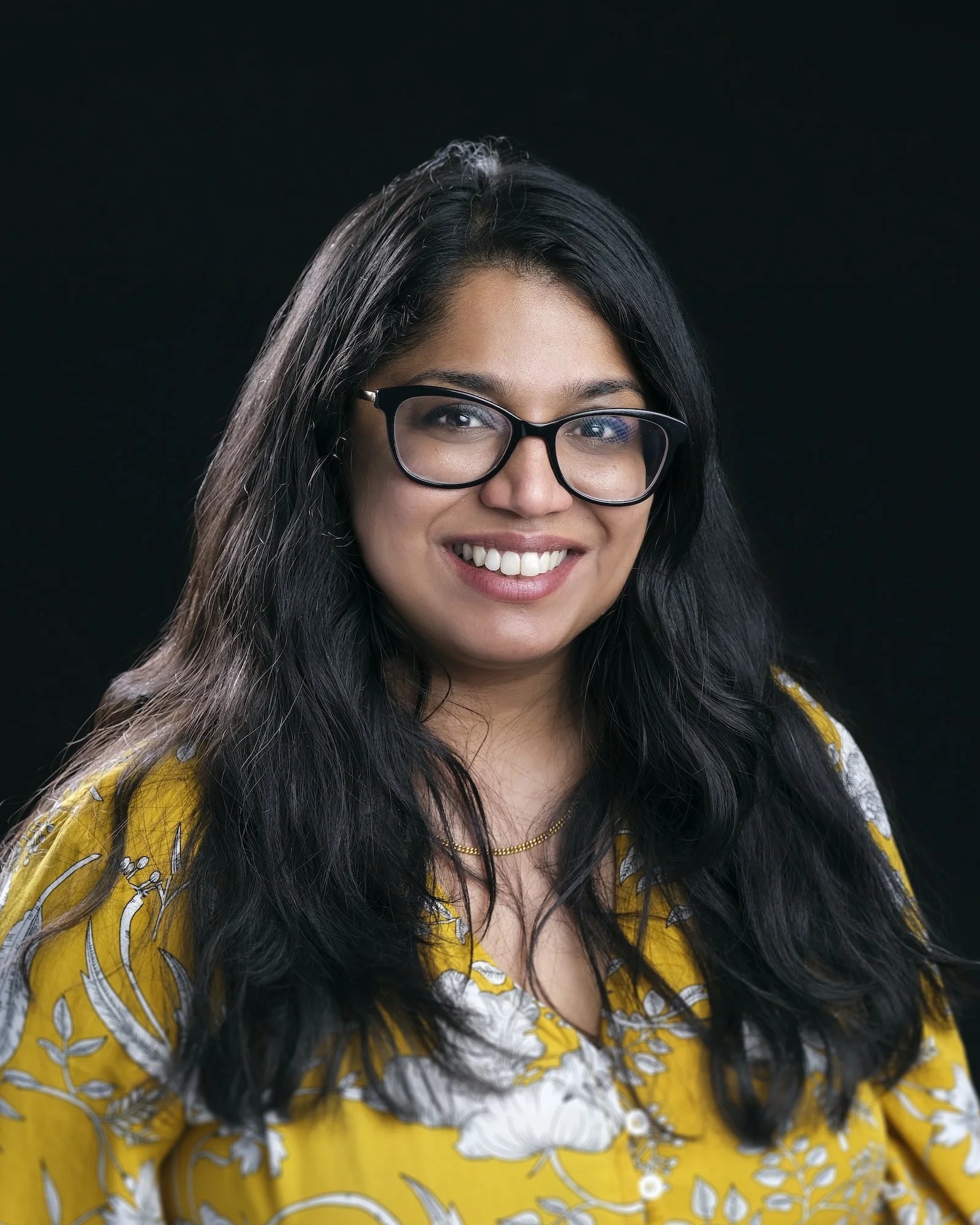 A woman with long black hair wearing black glasses and a yellow floral blouse, smiling against a black background.