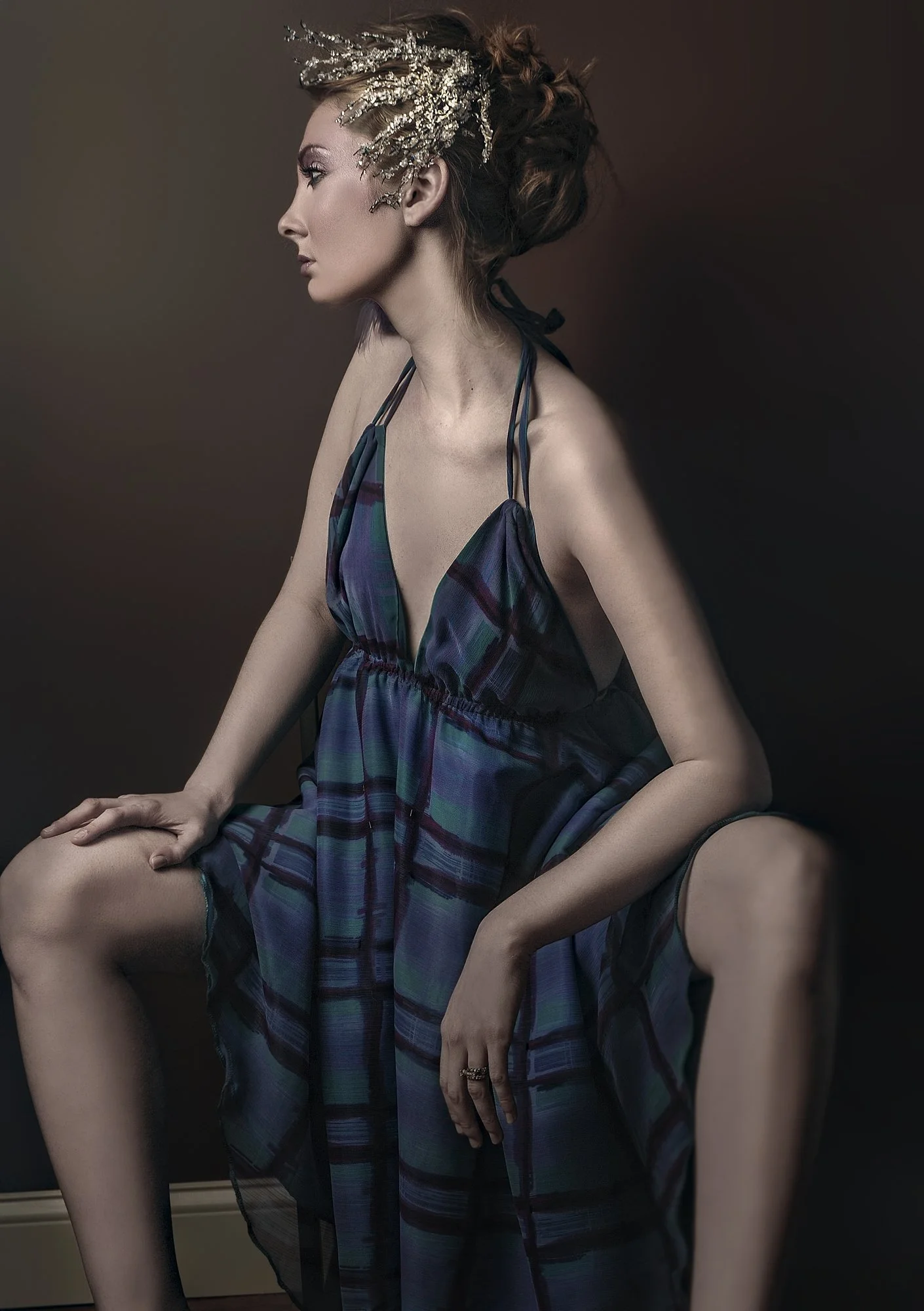 A woman in a plaid dress with thin straps, sitting with one knee up and one hand on her knee, wearing a decorative headpiece, against a dark background. Best-Fashion-Portrait-Commercial-Business-Headshots-Corporate- photography-Maryland-Washington D.