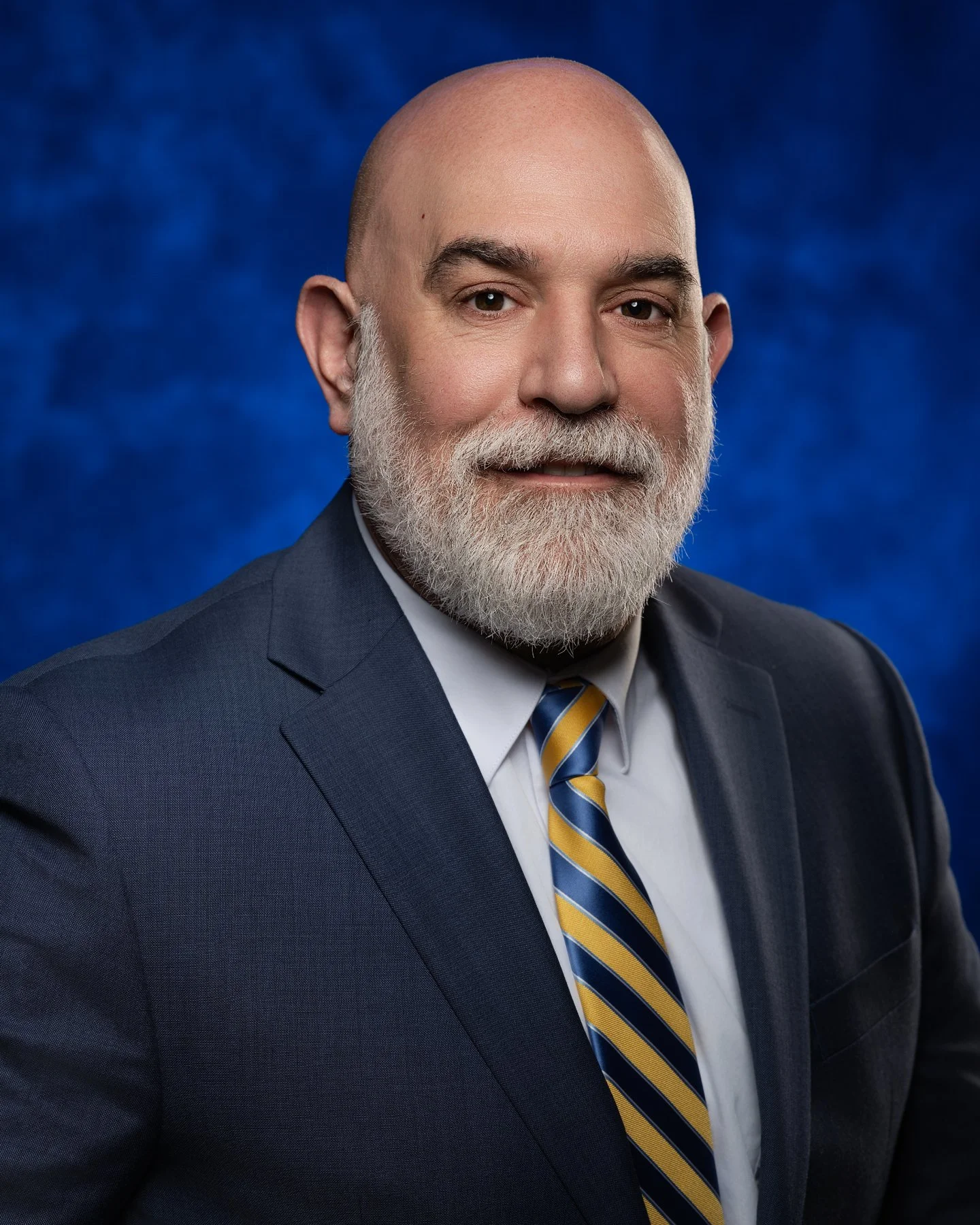 A professional headshot of a middle-aged man with a bald head, white beard, and mustache, wearing a dark suit, light dress shirt, and striped tie, against a blue textured background. In office headshot and branding photographer in Maryland, PA, VA an