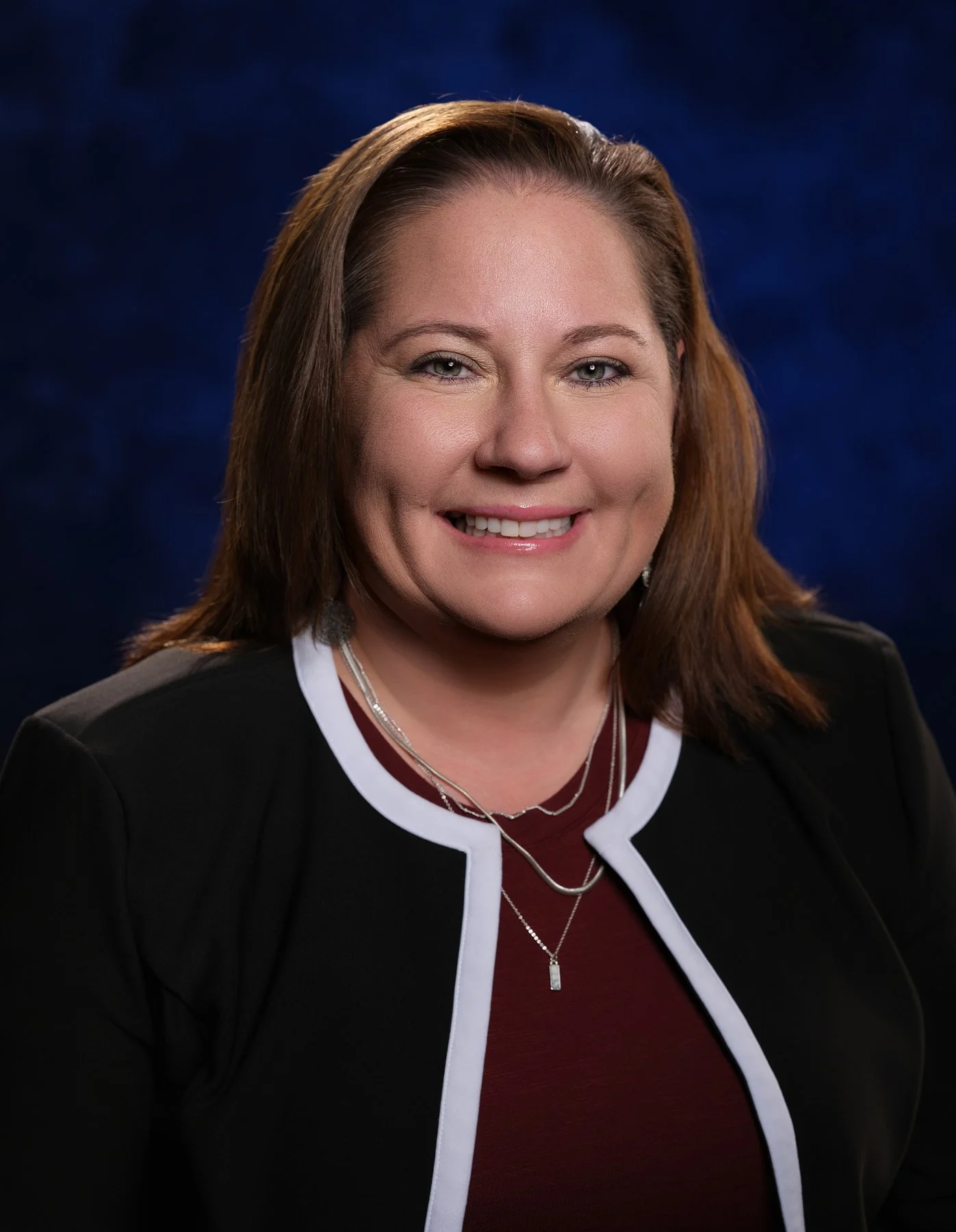 A smiling woman with shoulder-length brown hair, wearing a black blazer with white trim, a maroon top, and layered necklaces, posed against a dark blue background. In office headshot and branding photographer in Maryland, PA, VA and Washington D.C. b
