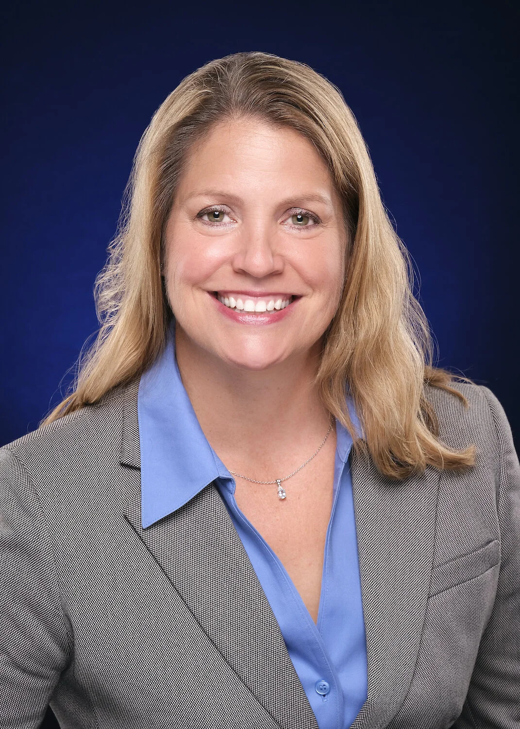 Professional headshot of a smiling woman with shoulder-length blonde hair, wearing a grey blazer over a blue shirt, necklace, against a dark blue background.