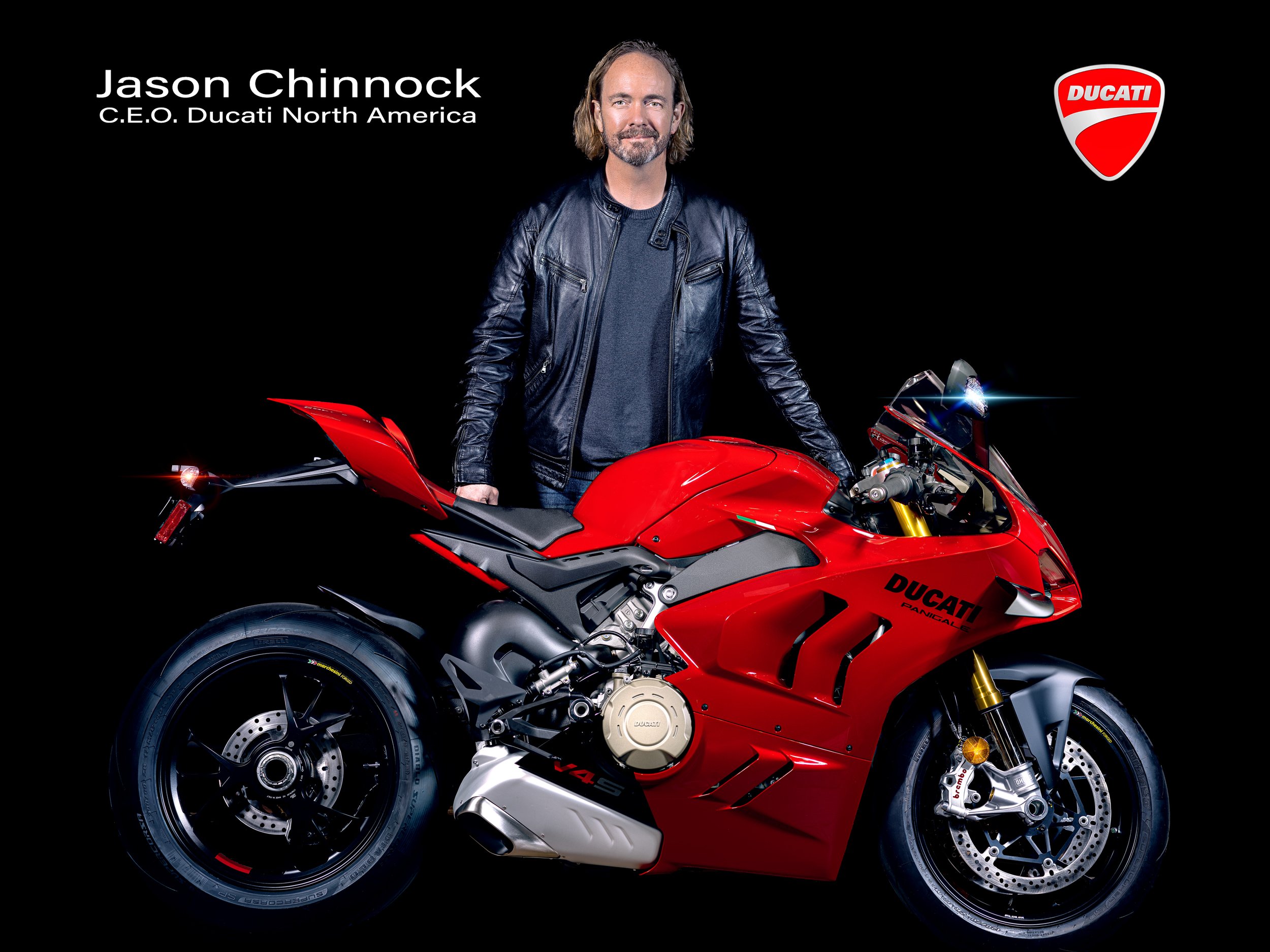 A man stands next to a red Ducati Panigale V4 motorcycle against a black background. The man is wearing a black leather jacket and has shoulder-length hair and a beard. The Ducati logo is visible on the bike and in the upper right corner of the image, and the man's name and title are on the top left corner.
