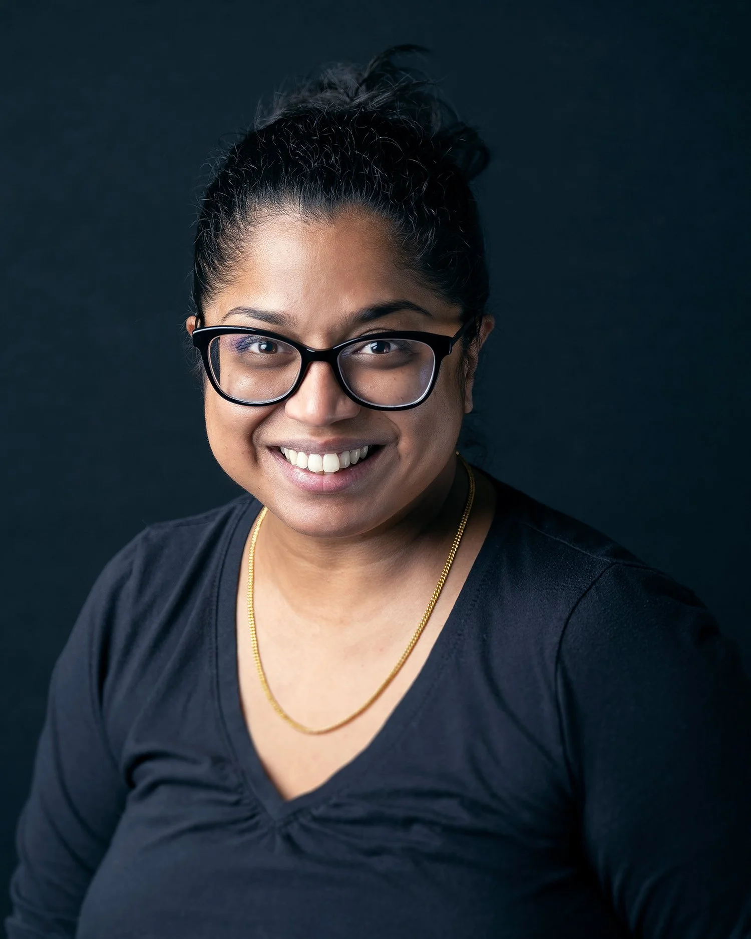 Close-up portrait of a smiling woman wearing glasses, a black shirt, a gold chain, with her hair in a bun against a dark background.