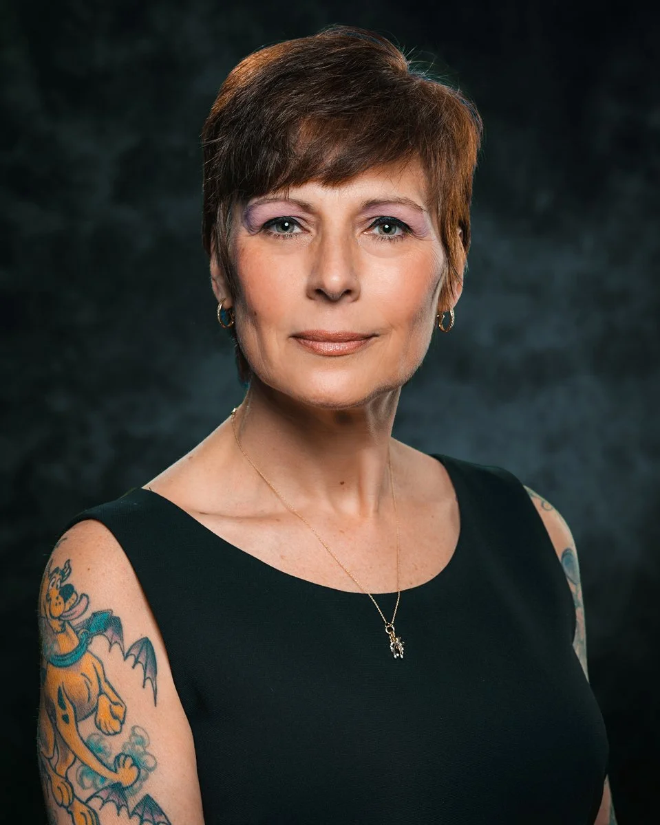 A portrait of a middle-aged woman with short brown hair, wearing a black sleeveless top, gold hoop earrings, a delicate gold necklace with a small pendant, and colorful tattoos on her arms, against a dark background.