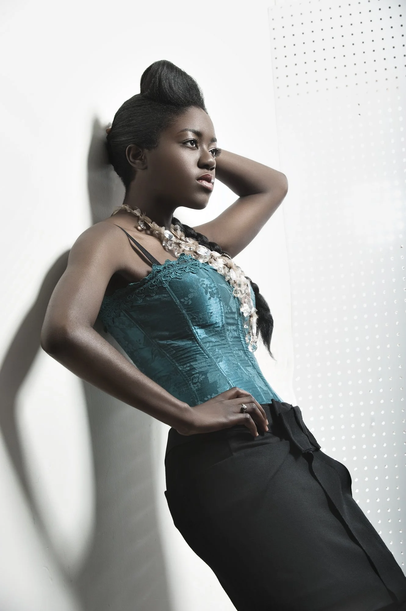 A woman with dark skin and long black braided hair, wearing a teal satin lace top and black high-waisted pants, posed with one hand on her hip and the other behind her head, leaning against a wall with a dotted light pattern. Best-Fashion-Portrait-Co