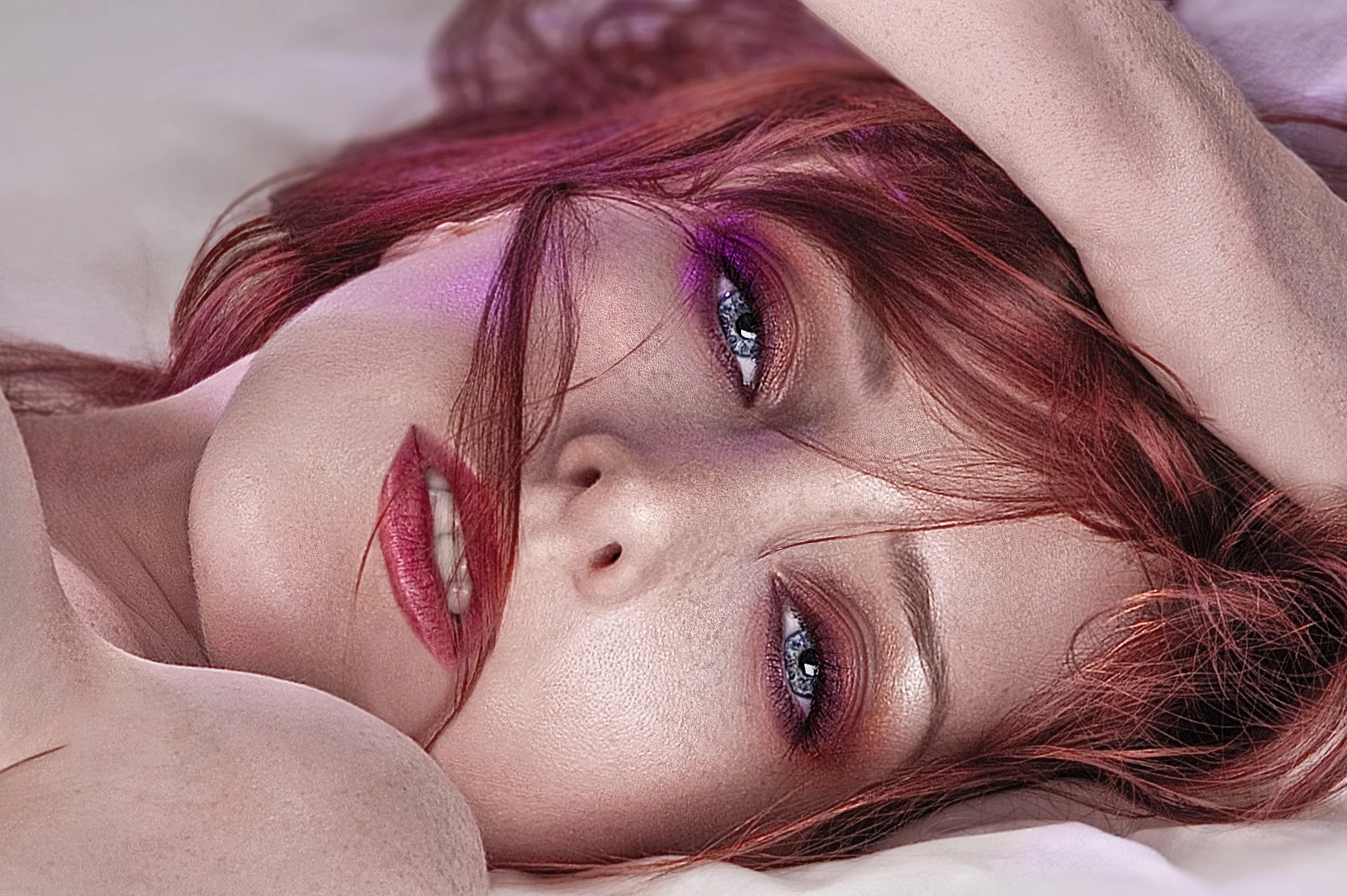 A close-up of a woman with red hair lying down, with her head resting on her arm, wearing pink and purple eye makeup and pink lipstick.  Dynamic fashion branding and portrait photography by www.exvotostudio.com located in Maryland U.S.A. 