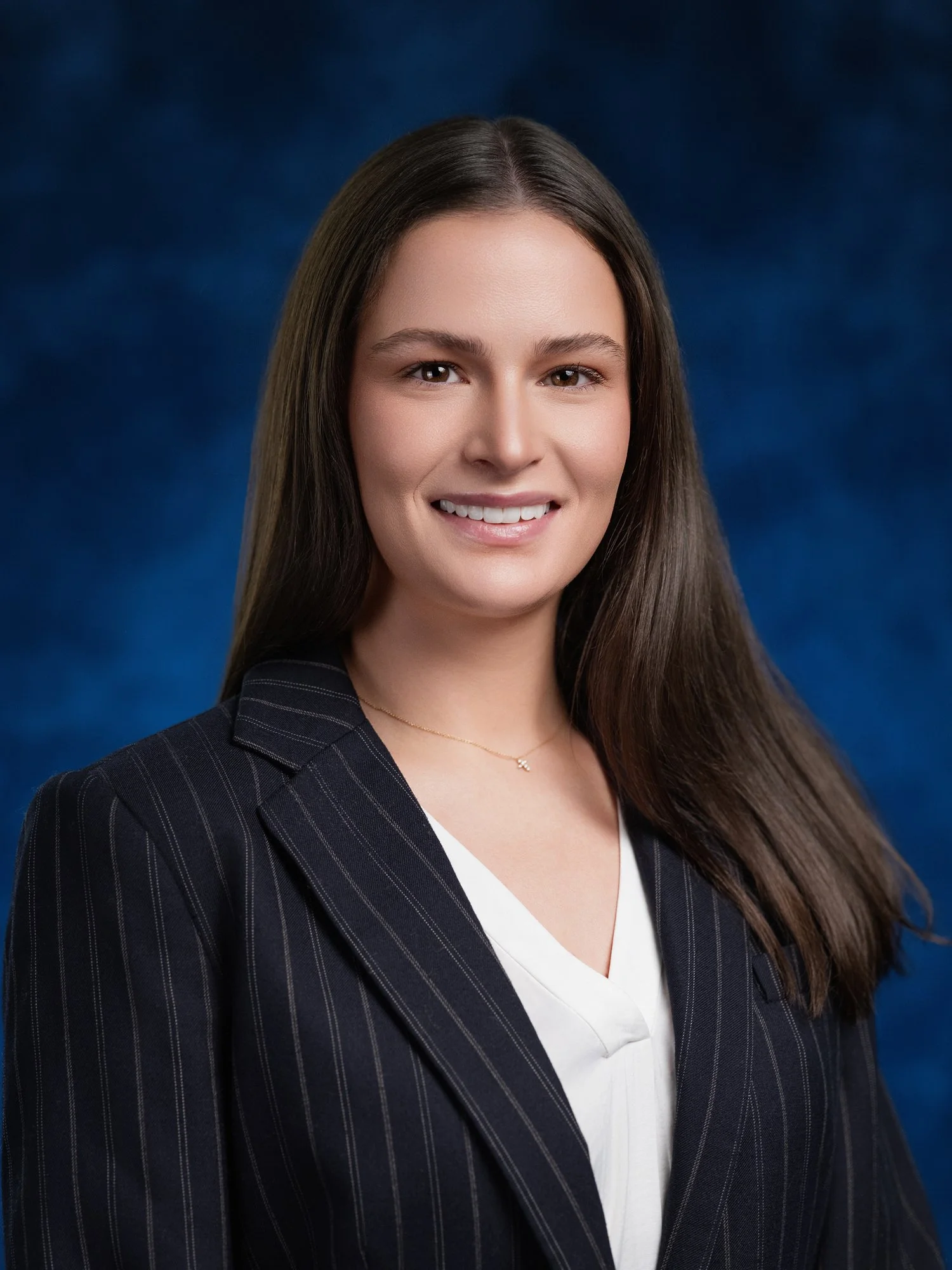 Professional portrait of a woman with long brown hair, wearing a dark striped blazer and a white blouse, smiling against a blue background. In office headshot and branding photographer in Maryland, PA, VA and Washington D.C. by www.exvotostudio.com