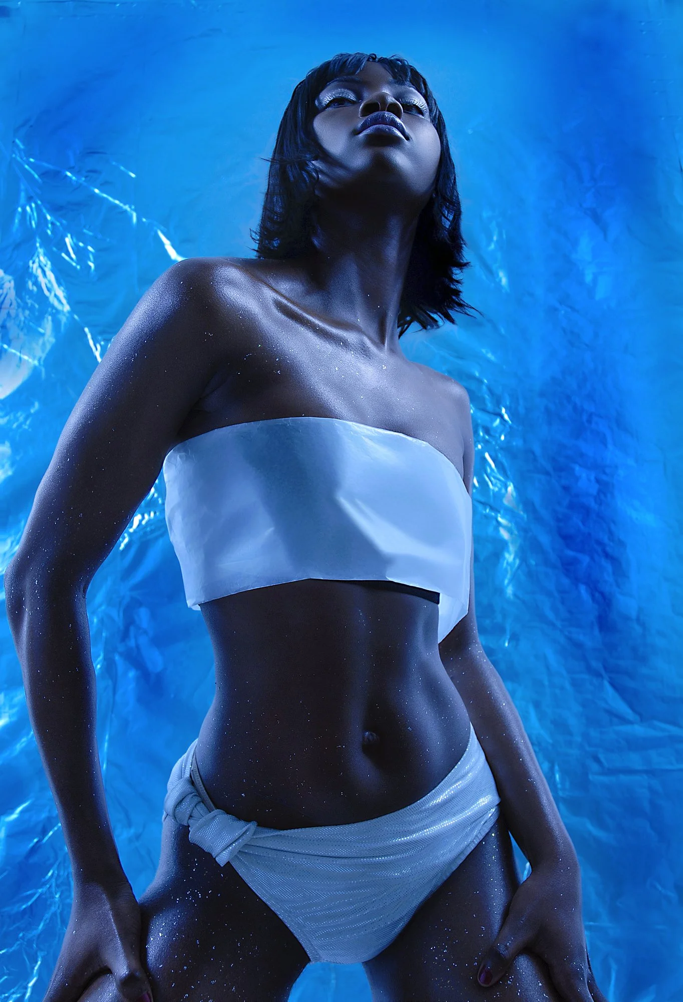 A woman with short dark hair and shiny dark lipstick poses with her head tilted upward against a crinkled, glowing blue background. She is wearing a white strapless top and matching shorts, with glitter on her skin. Best-Fashion-Portrait-Commercial-B