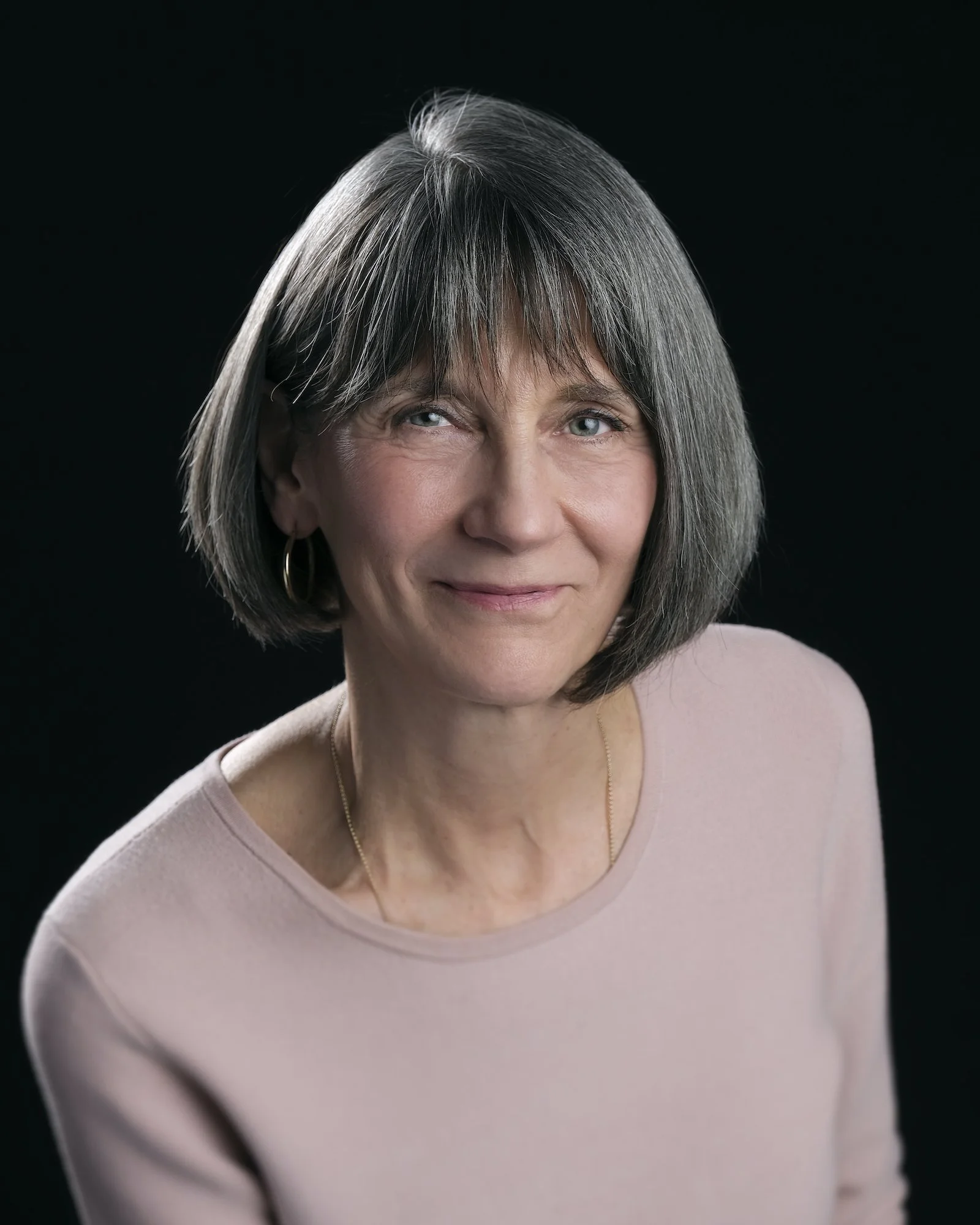 A woman with short gray hair and blue eyes, wearing a light pink top and gold jewelry, smiling softly against a dark background.
