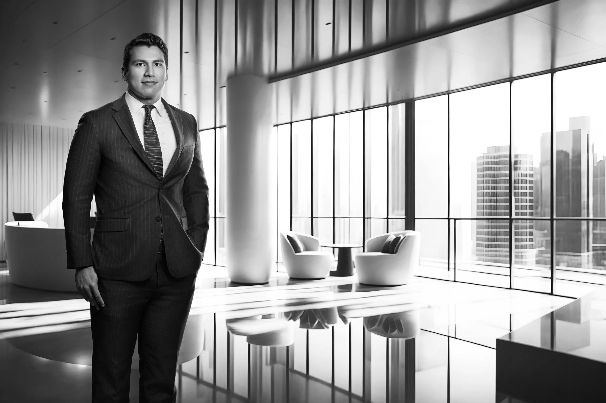 A man in a dark suit standing in a modern office lobby with large windows, minimal furniture, and city buildings in the background. www.exvotostudio.com
