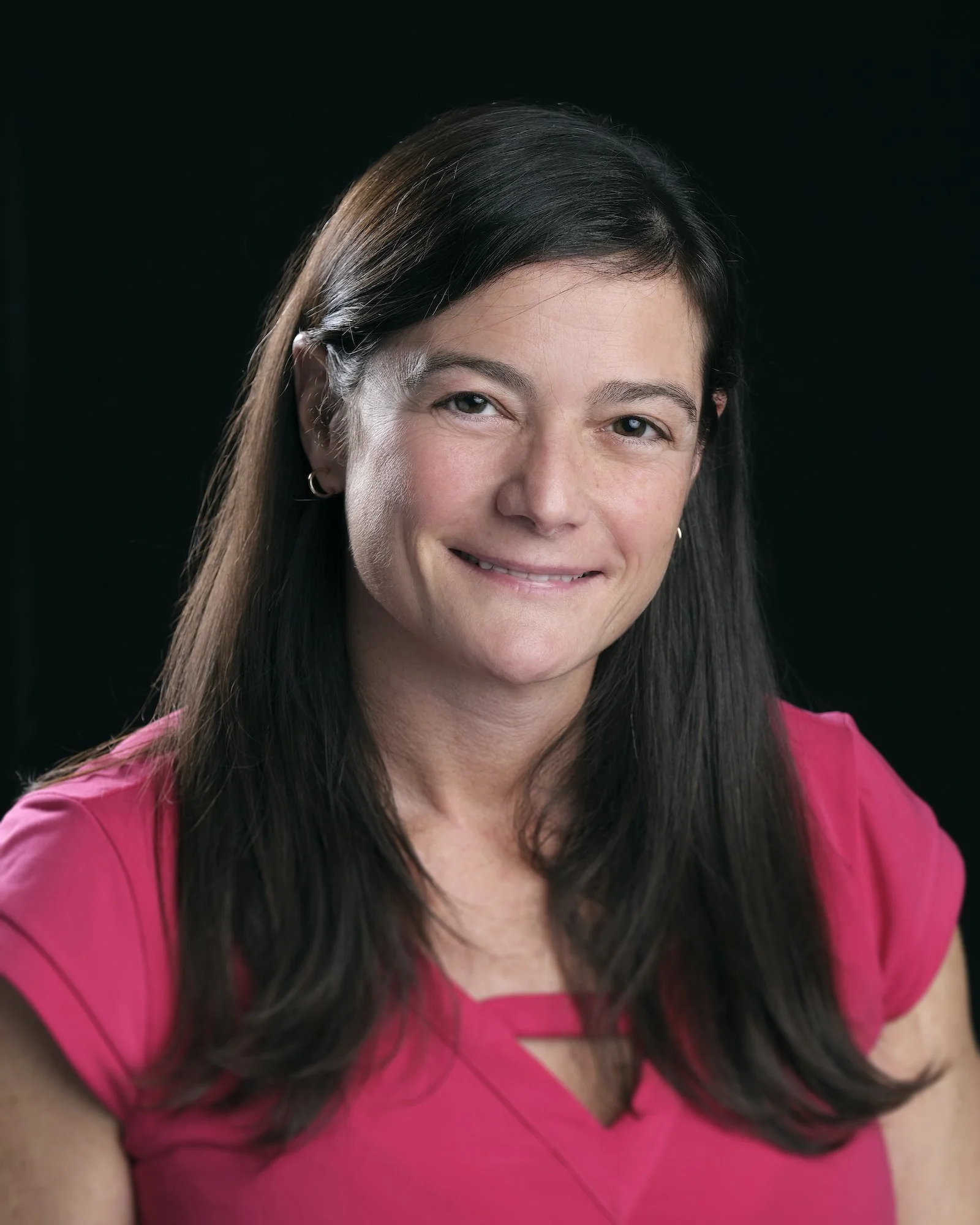 Portrait of a woman with dark hair, smiling, wearing a pink top, against a black background.