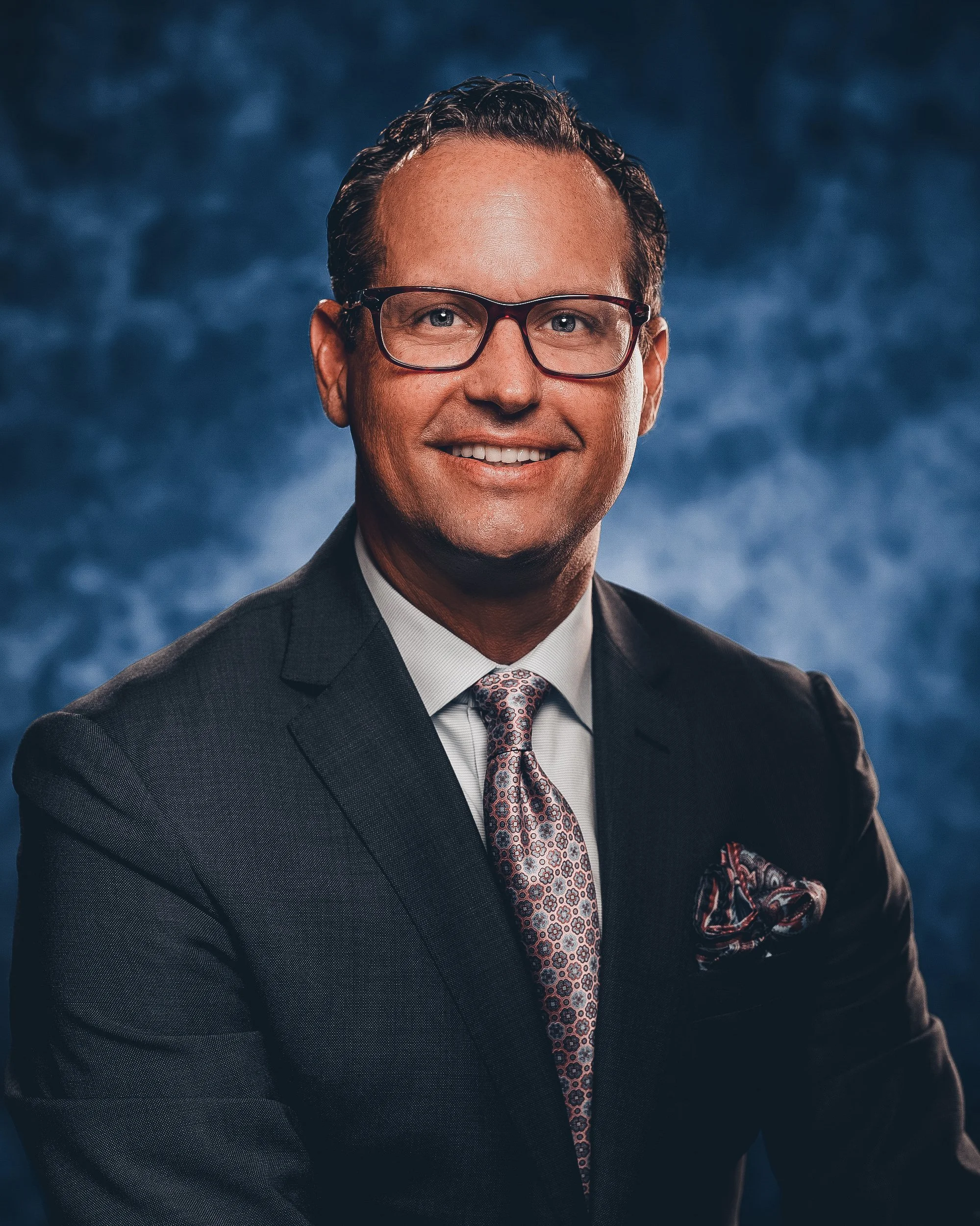 A professional portrait of a man in a suit and tie, wearing glasses, smiling against a blue background.