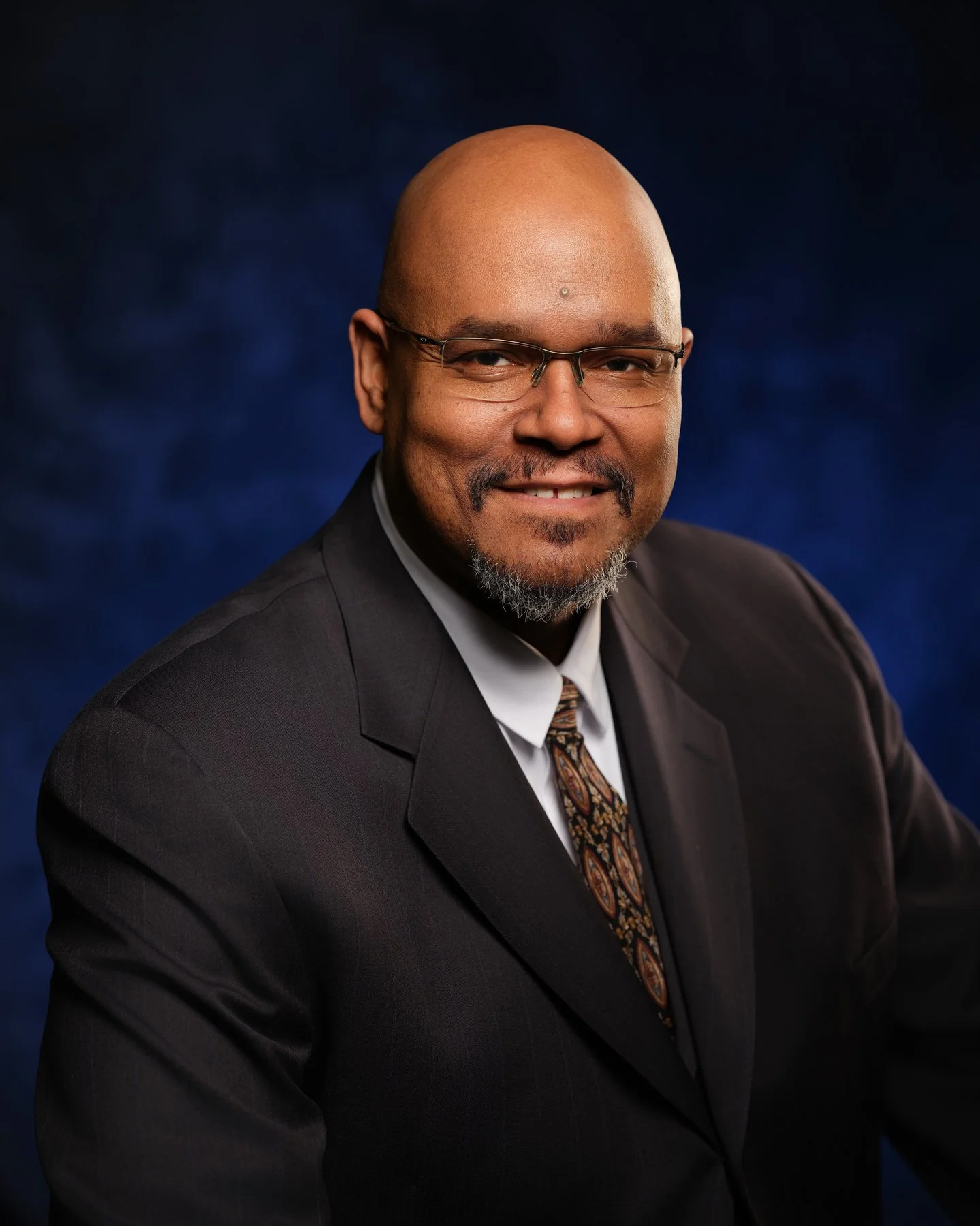 Professional portrait of a bald man with glasses, wearing a dark suit, white shirt, and patterned tie, smiling against a dark blue background. In office headshot and branding photographer in Maryland, PA, VA and Washington D.C. by www.exvotostudio.co
