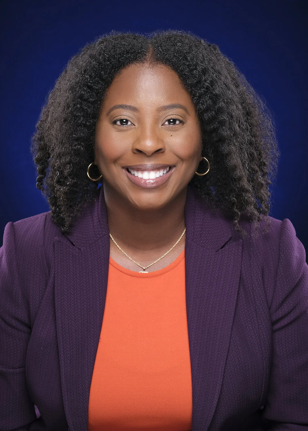 A smiling woman with dark curly hair wearing a purple blazer and orange top, against a dark blue background.
