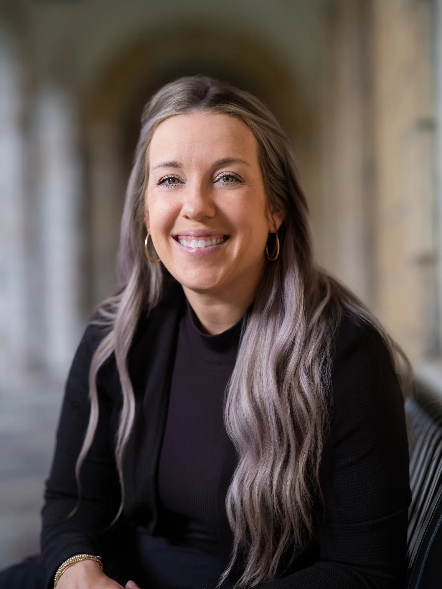 Portrait of a smiling woman with long wavy gray hair, wearing gold hoop earrings and a black top, seated outdoors with a blurred architectural background.