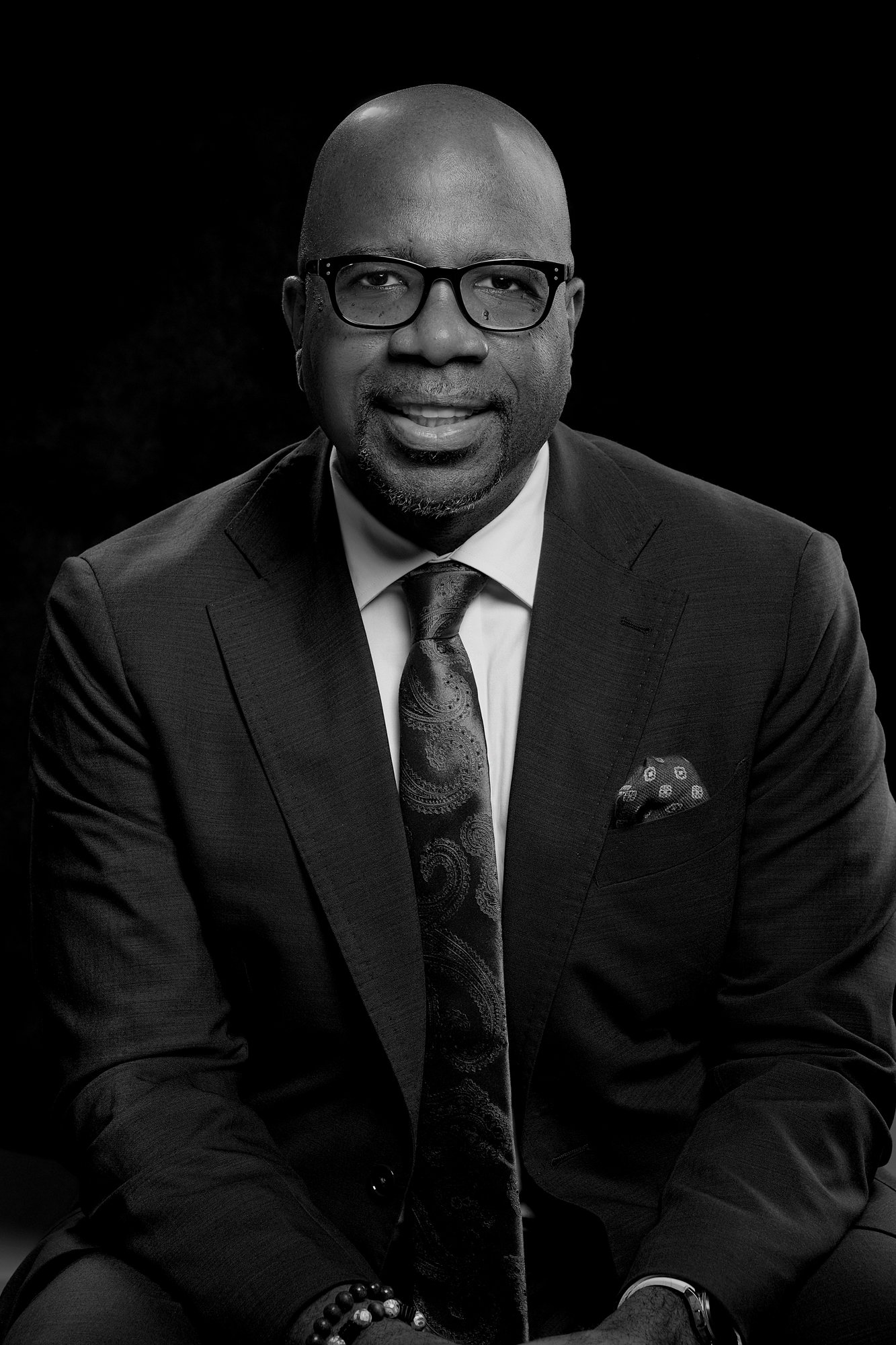 Black and white portrait of a man wearing glasses, a dark suit, a patterned tie, and a matching pocket square, sitting against a dark background.