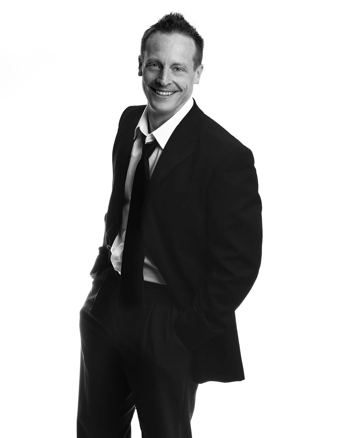 Black and white portrait of a smiling man in a suit with hands in pockets, looking at the camera.