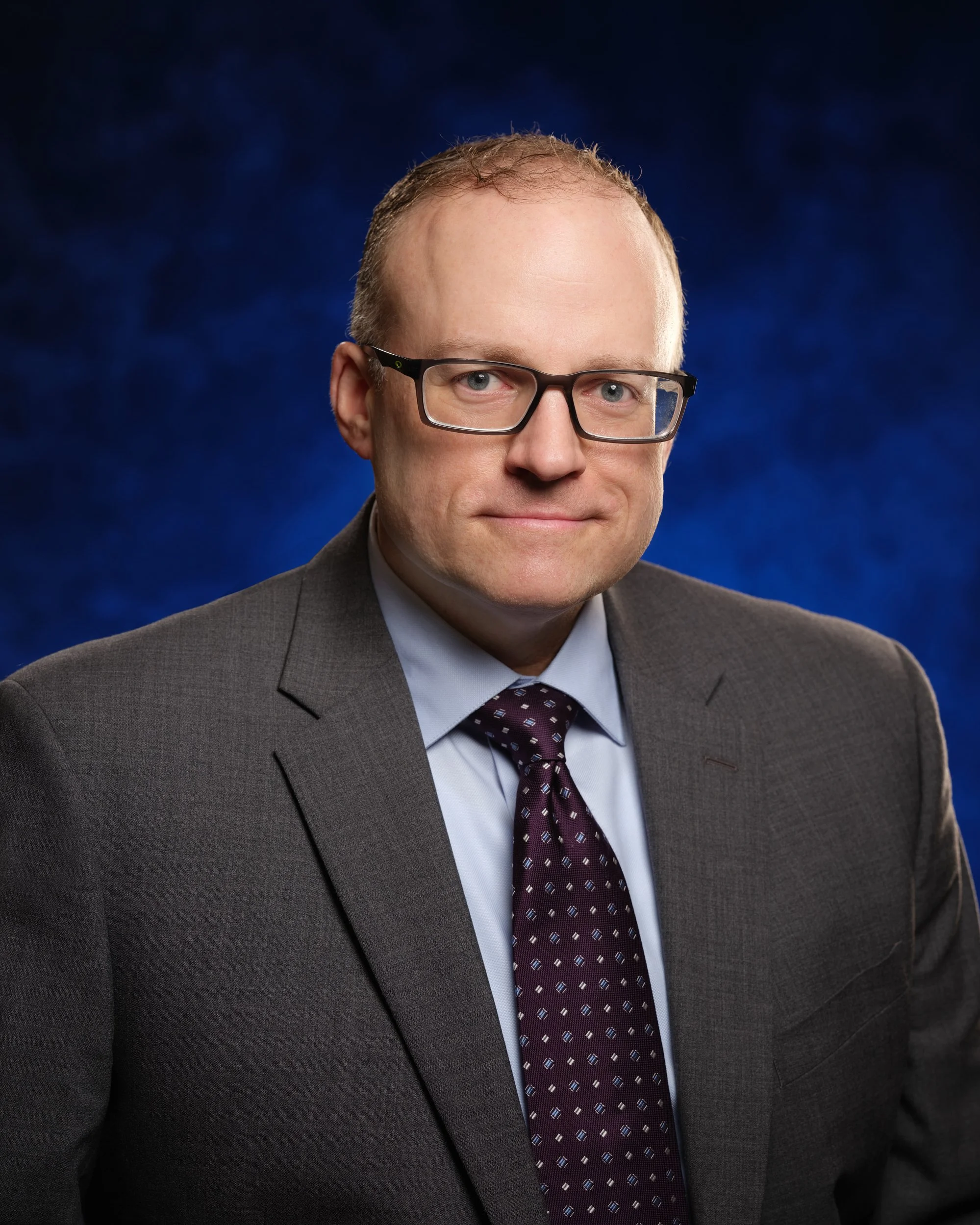 Headshot of a man in a suit and tie, wearing glasses, against a dark blue background.