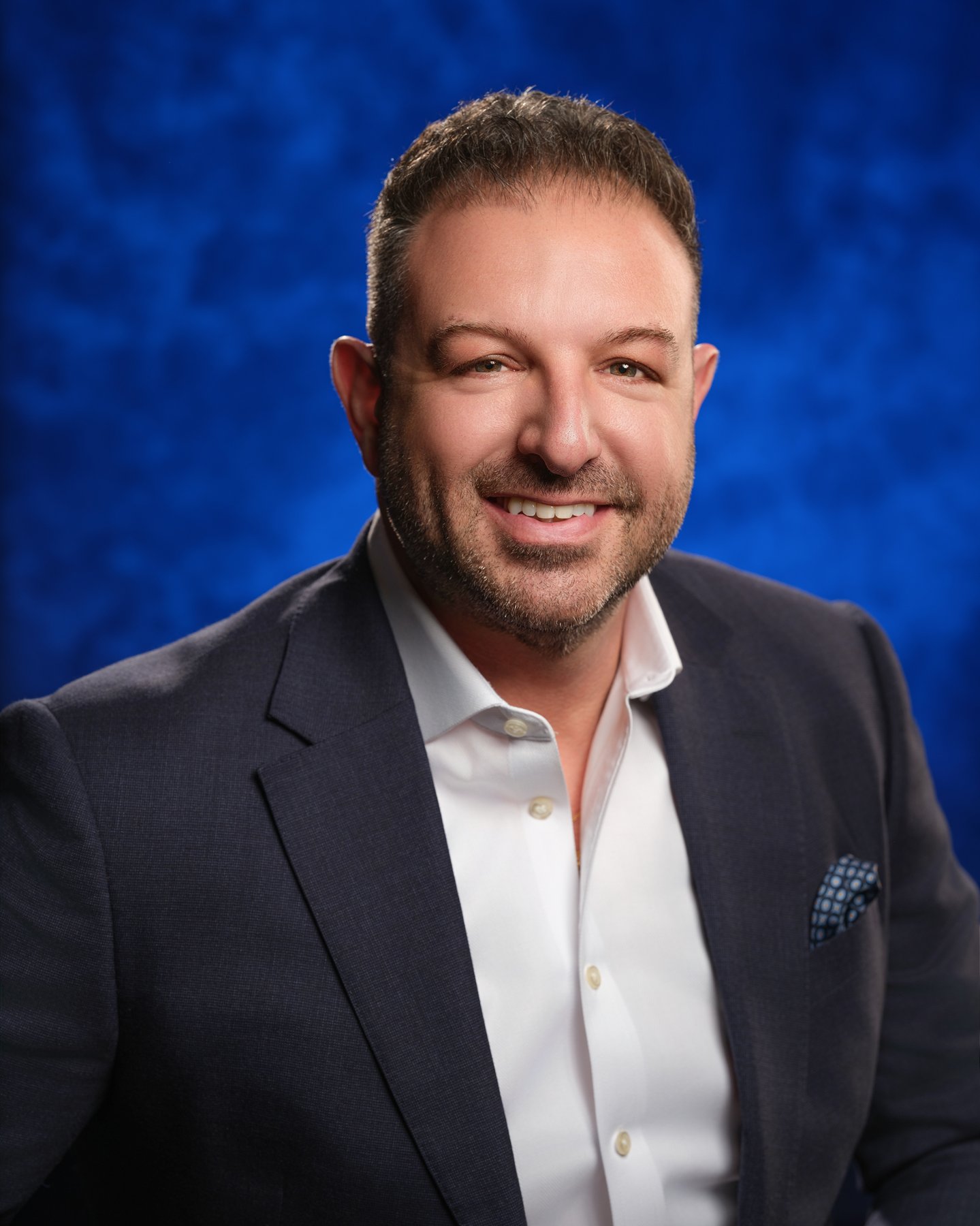 A man with short brown hair, a beard, and a smile, wearing a dark suit jacket, white dress shirt, and a checkered pocket square, against a blue textured background. In office headshot and branding photographer in Maryland, PA, VA and Washington D.C. 