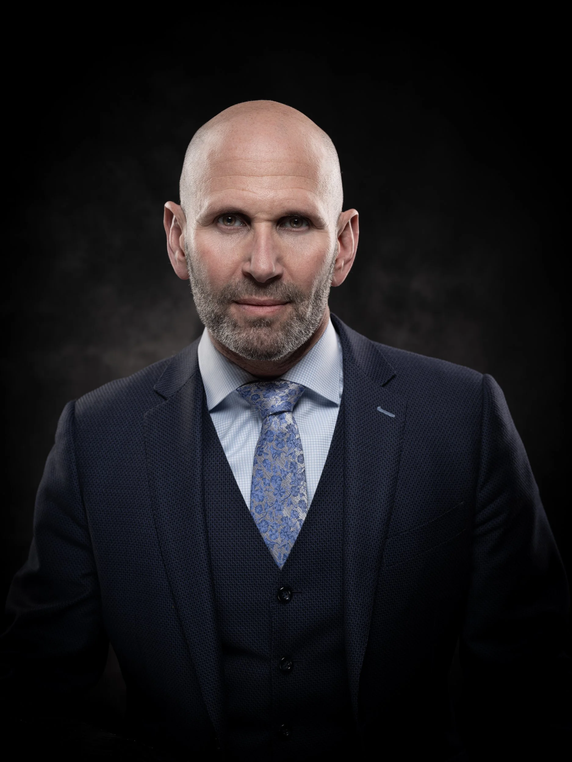 A bald, middle-aged man with a beard wearing a dark suit, light blue shirt, and patterned tie, standing against a dark background.