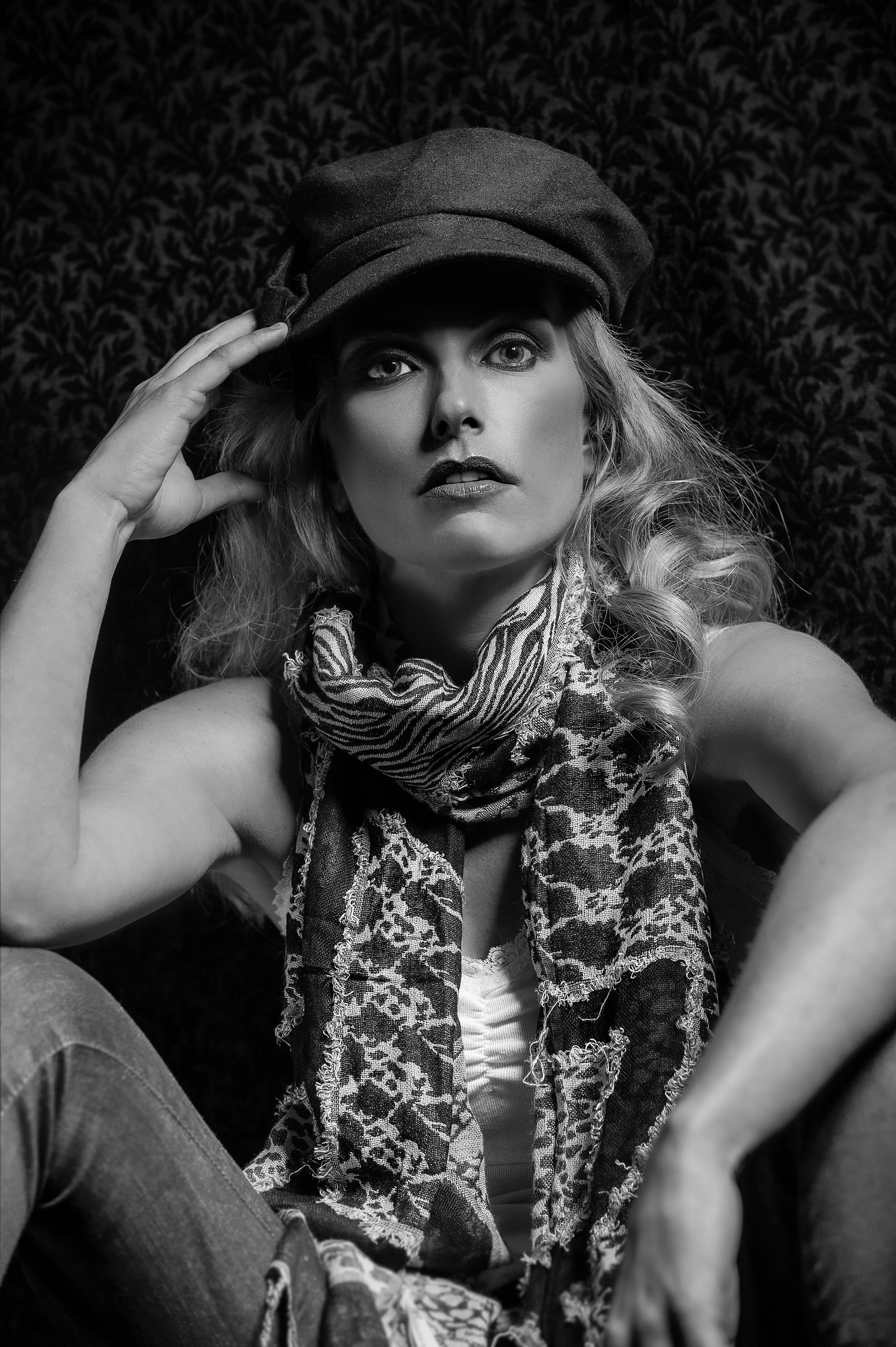 Black and white portrait of a woman with wavy hair, wearing a newsboy cap, a patterned scarf, and casual clothing, sitting against a dark patterned background.