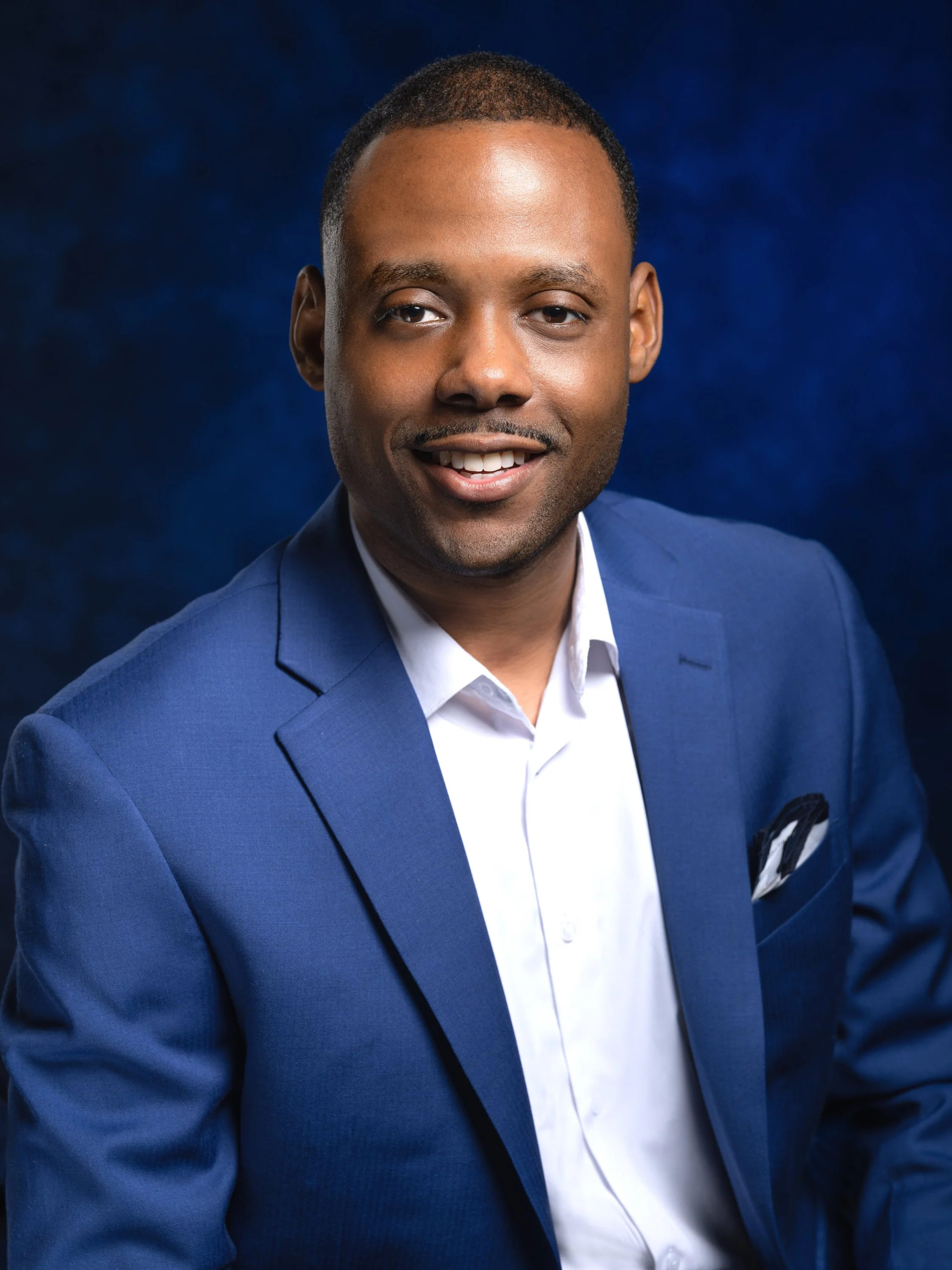 Professional headshot of a smiling Black man wearing a blue suit and white shirt against a dark blue background.