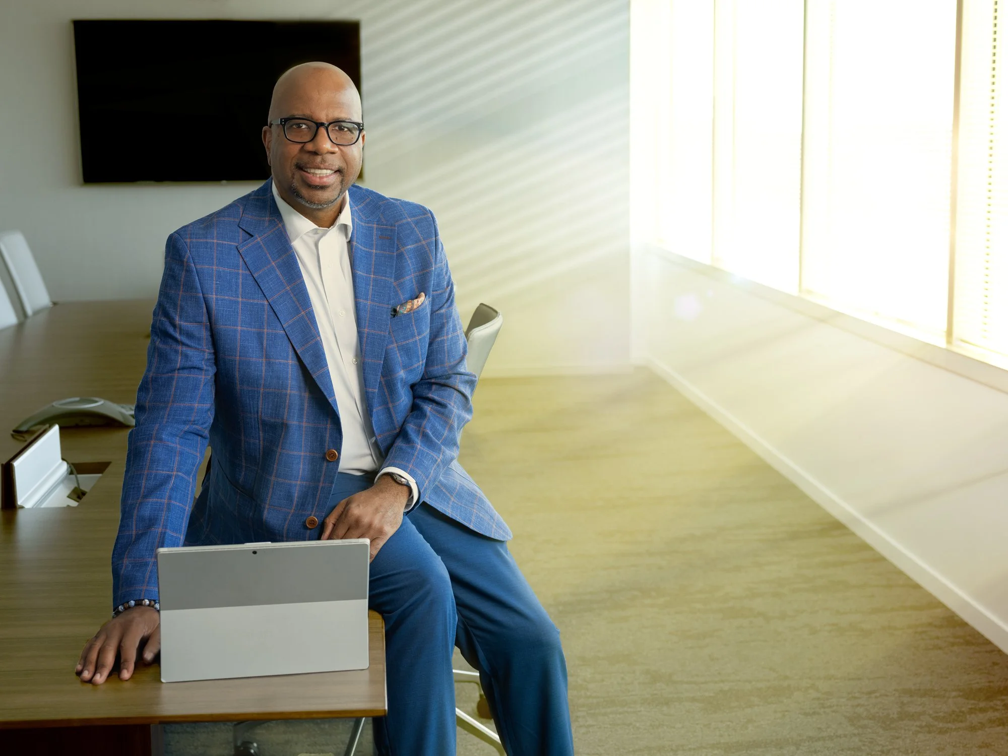 A man in a blue checkered suit and glasses sits on the edge of a conference table in a well-lit modern office, smiling at the camera with a tablet in front of him, and a TV screen on the wall behind him.