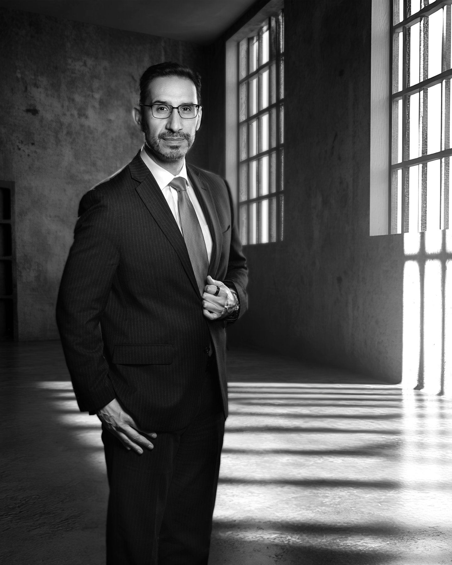 Black and white photo of a man in a suit standing in an industrial-style room with large windows, sunlight casting shadows on the floor.