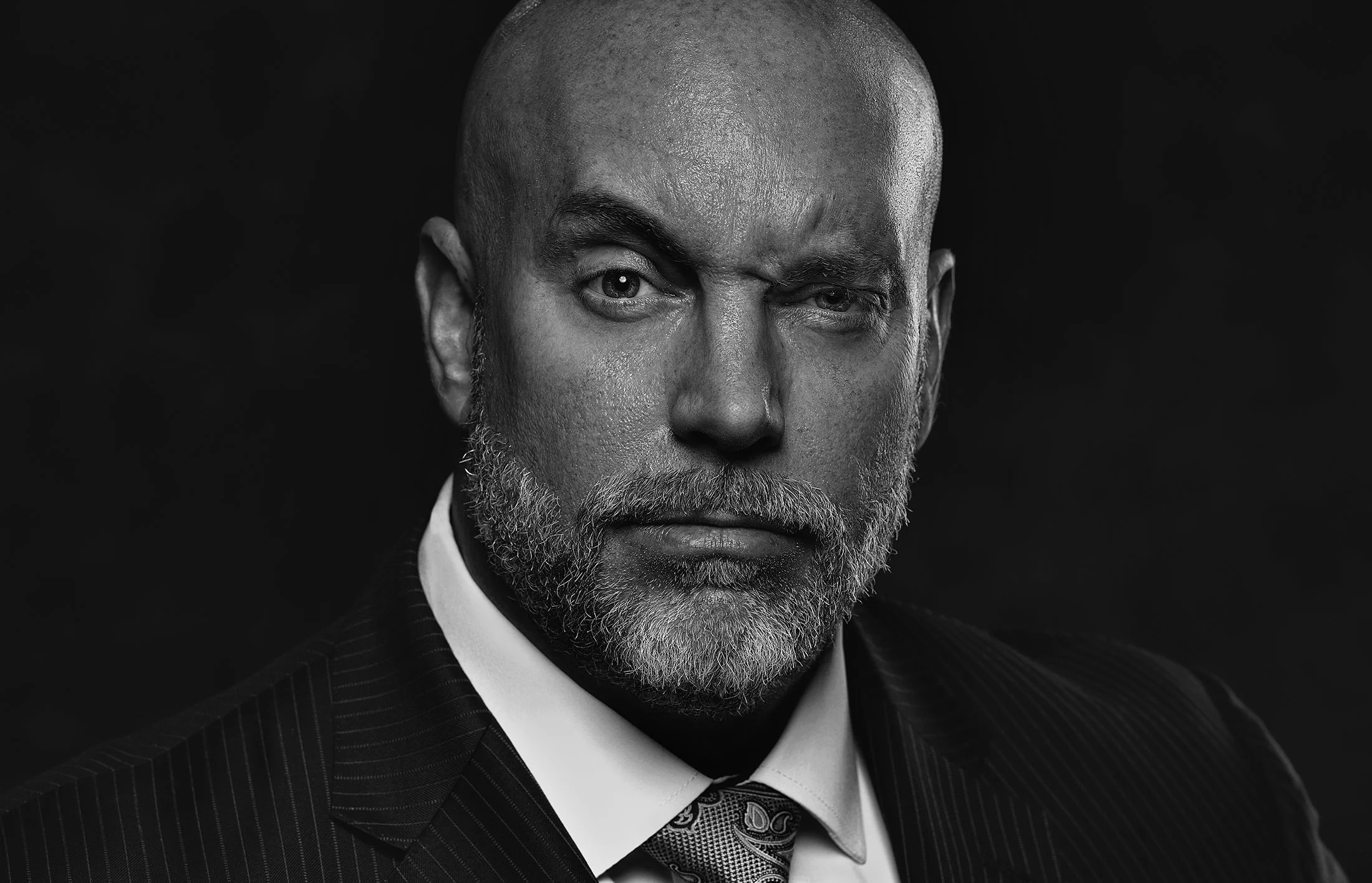 A black and white portrait of a middle-aged man with a bald head and gray beard, wearing a suit and tie, looking directly at the camera with one eye slightly squinted.