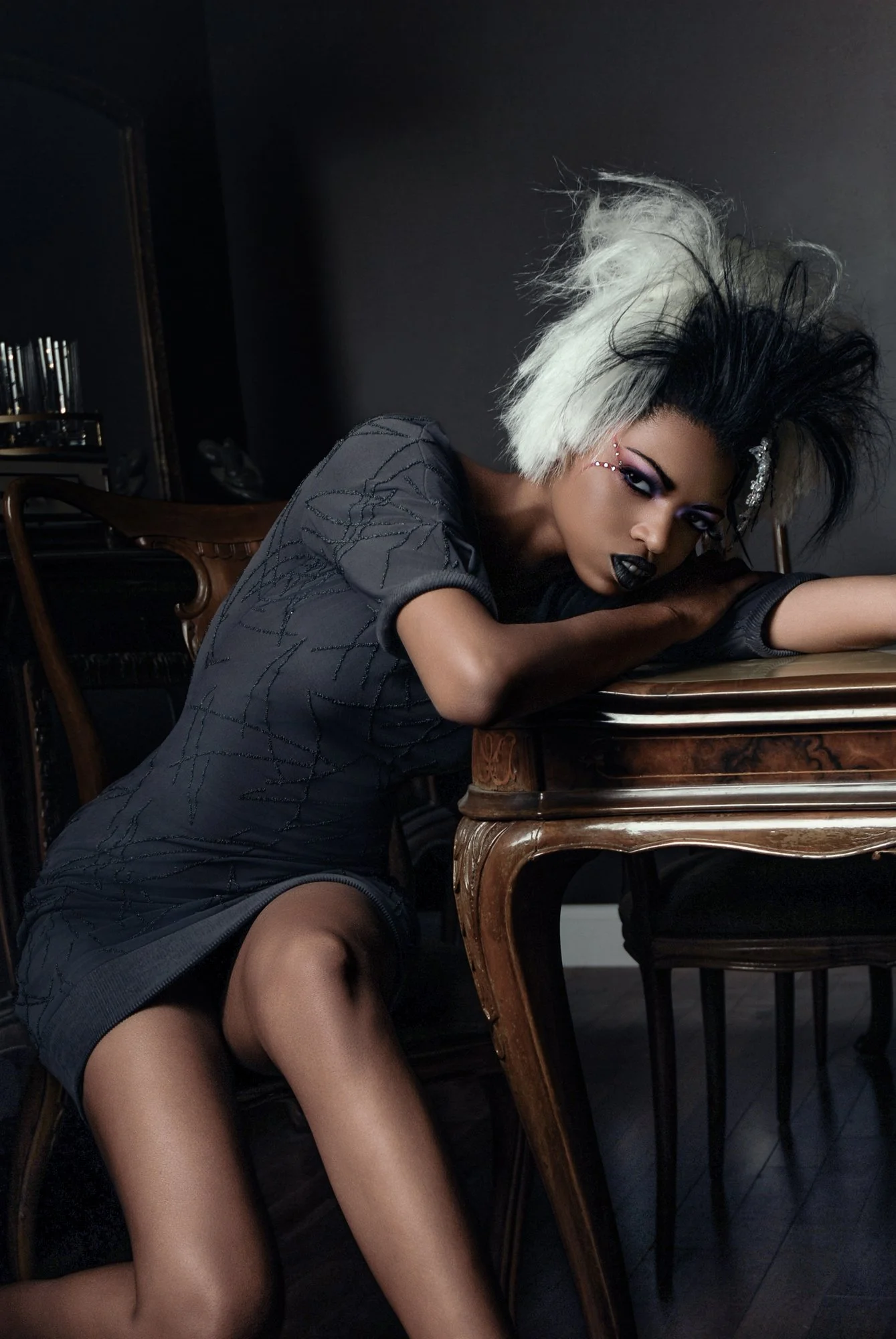 A woman with black and white hair, dark makeup, and black lipstick, leaning on a wooden dining table in a dark room with black walls, wearing a dark, textured dress. Best-Fashion-Portrait-Commercial-Business-Headshots-Corporate- photography-Maryland-