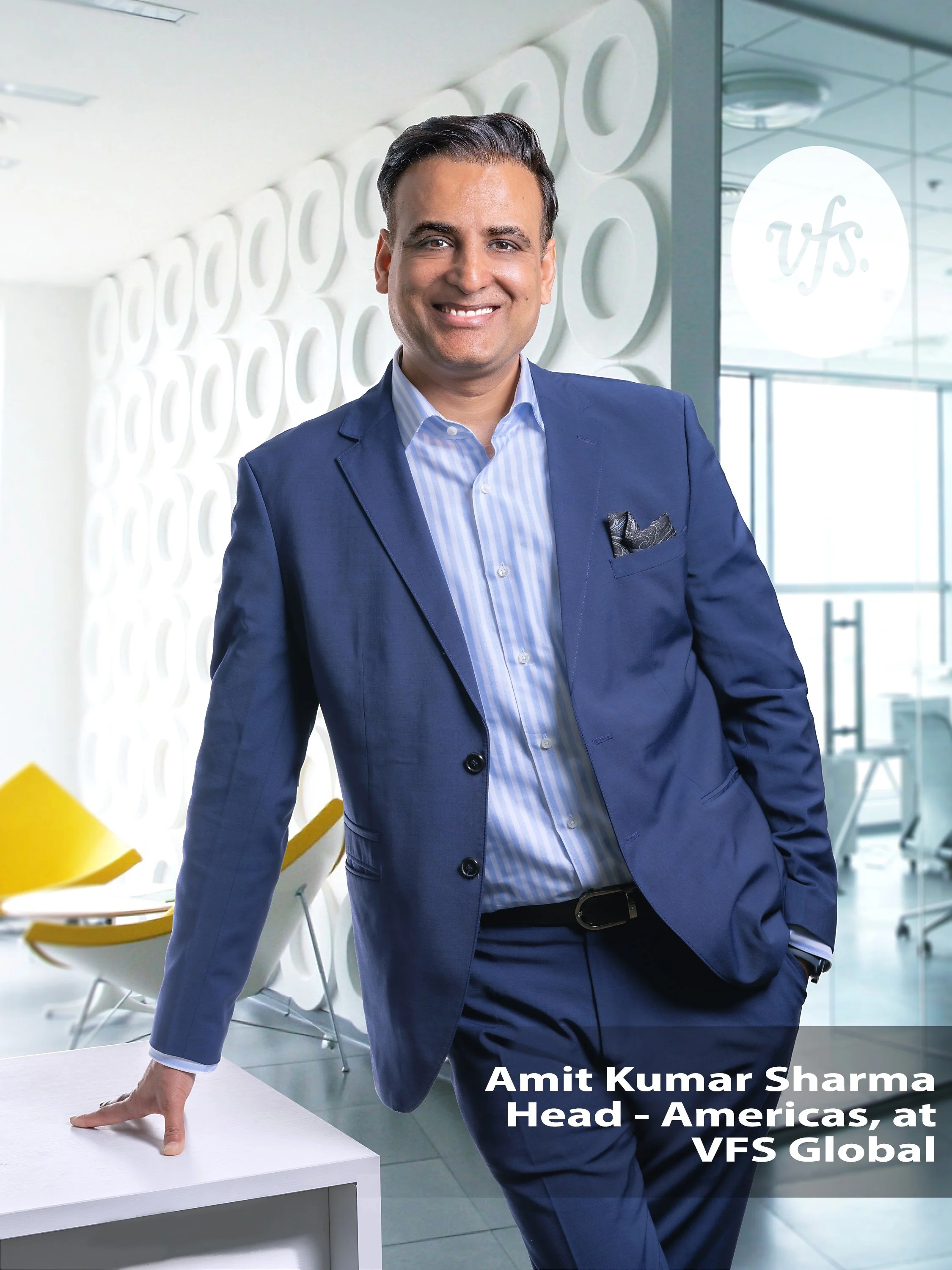 Man in a blue suit smiling, standing in a modern office with white geometric wall design, hand on desk. Text overlay reads "Amit Kumar Sharma, Head - Americas, at VFS Global." Yellow chairs in the background.