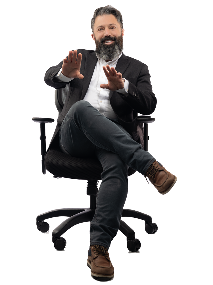 A man with a beard and gray hair, dressed in a dark blazer, white shirt, and jeans, sitting in an office chair with his legs crossed, smiling, and holding his hands up as if gesturing.