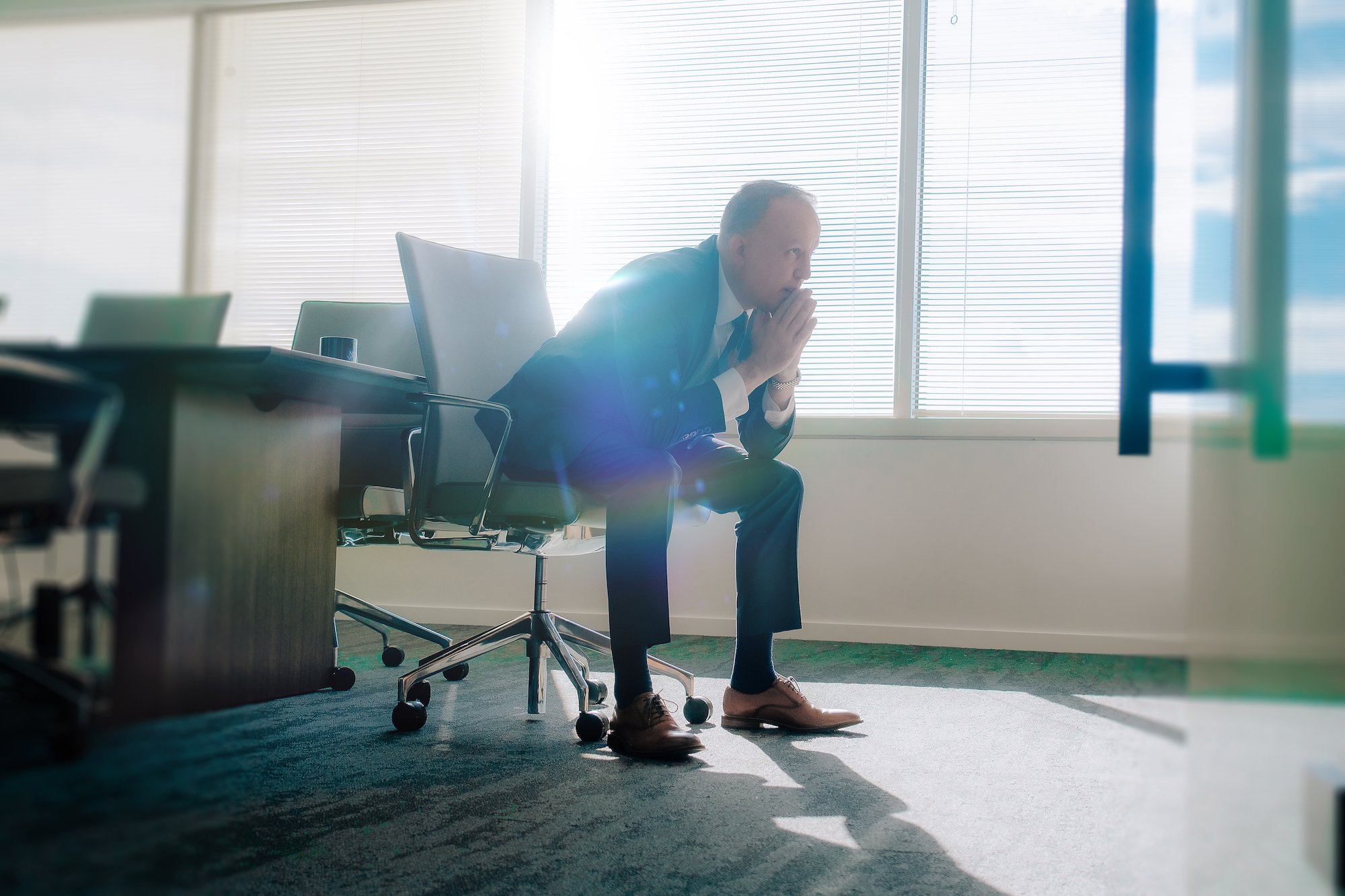 A man in a suit sitting on an office chair with his hands clasped in front of his face, appearing thoughtful or stressed, in a bright office with large windows and blinds.