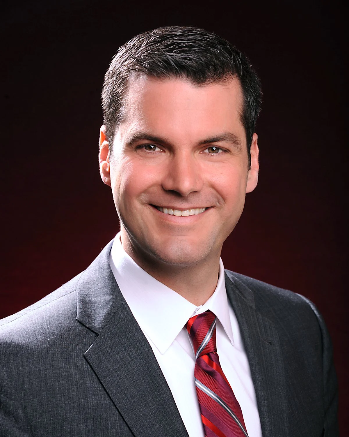 Headshot of a man smiling, wearing a dark suit, white shirt, and red striped tie, against a dark background. In office headshot and branding photographer in Maryland, PA, VA and Washington D.C. by www.exvotostudio.com